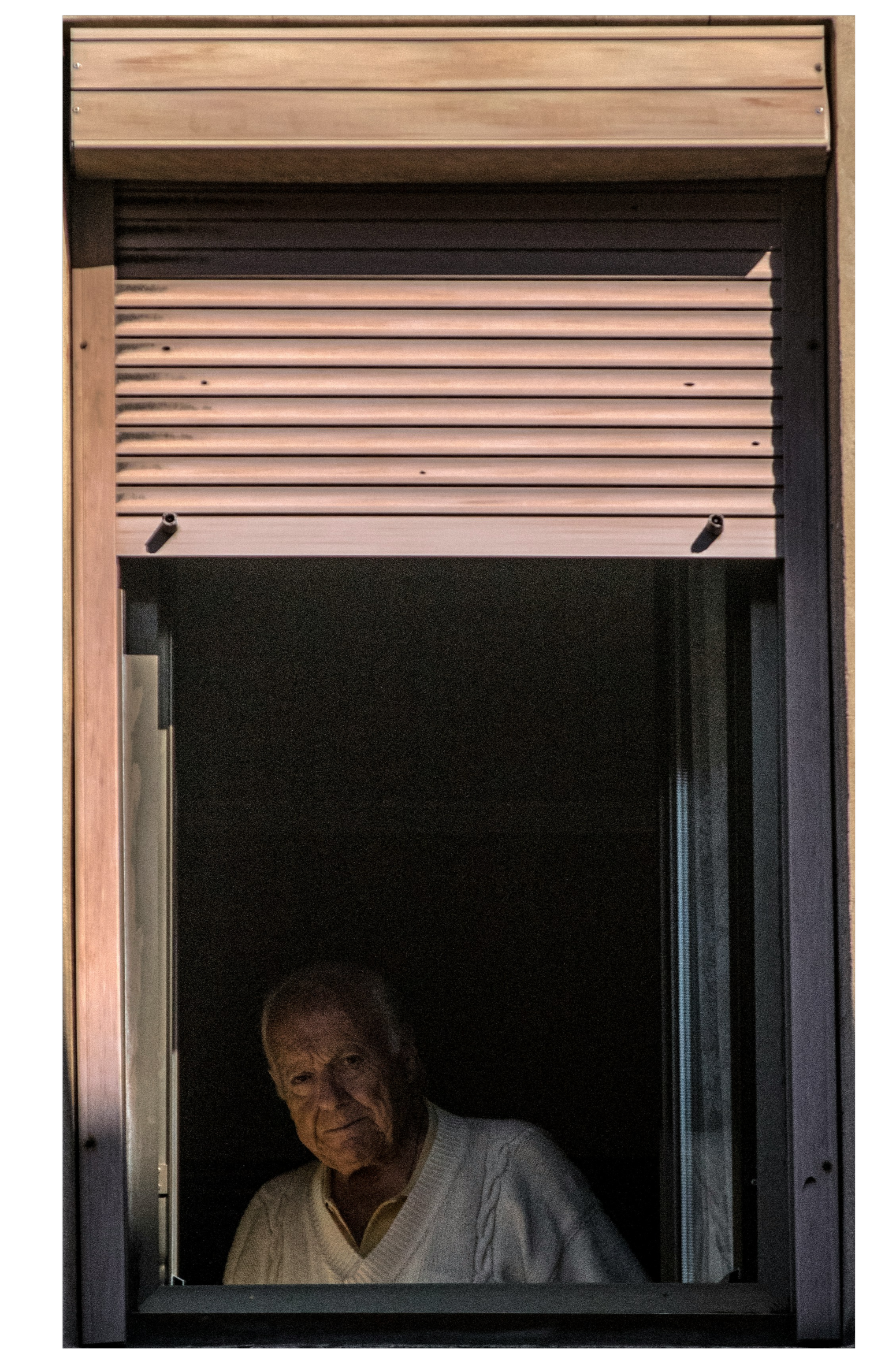 An elderly man standing at a window with a partially closed metal shutter, looking outside.