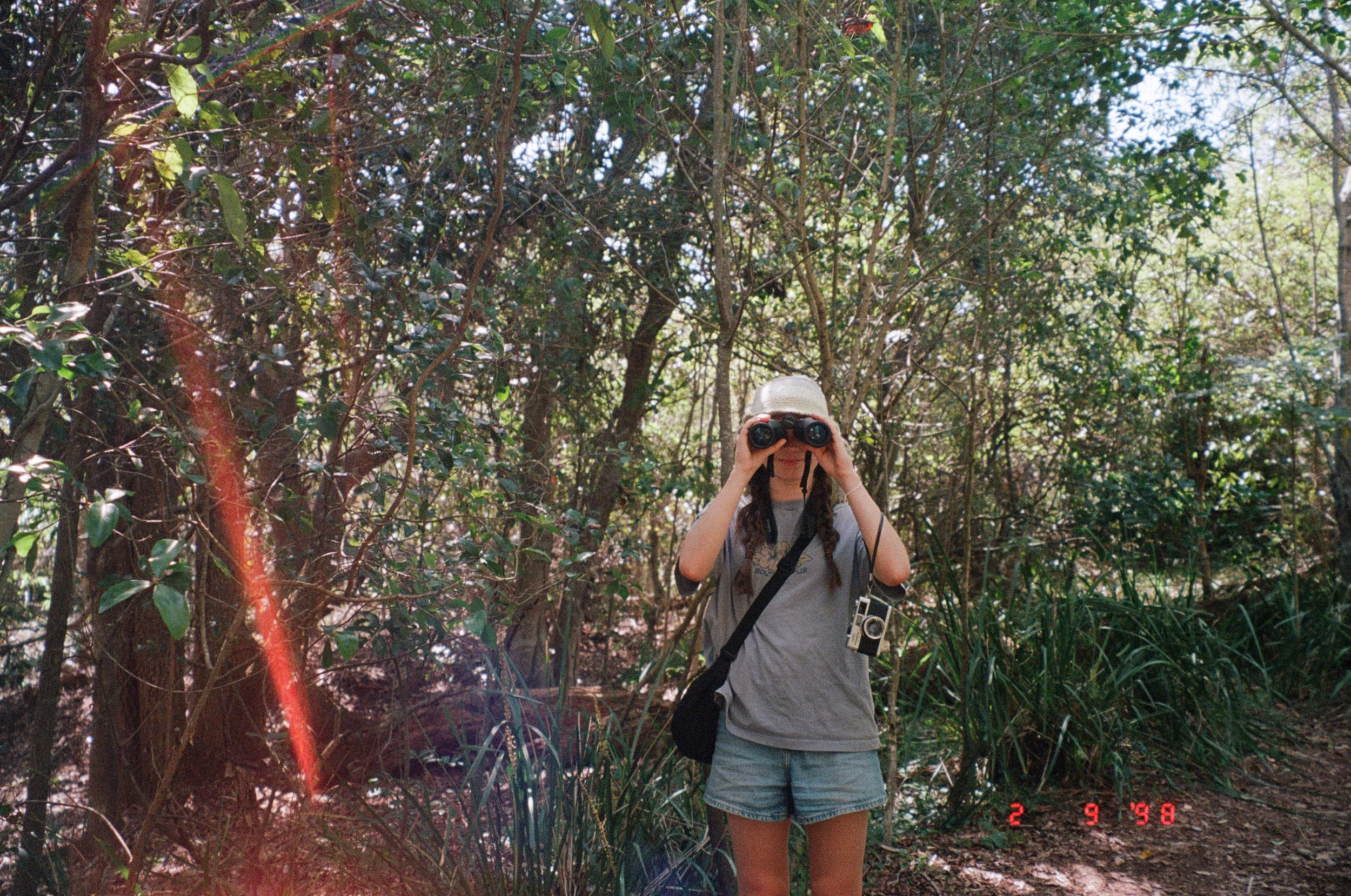 Person wearing a white hat and gray t-shirt looking through binoculars in a dense forest.