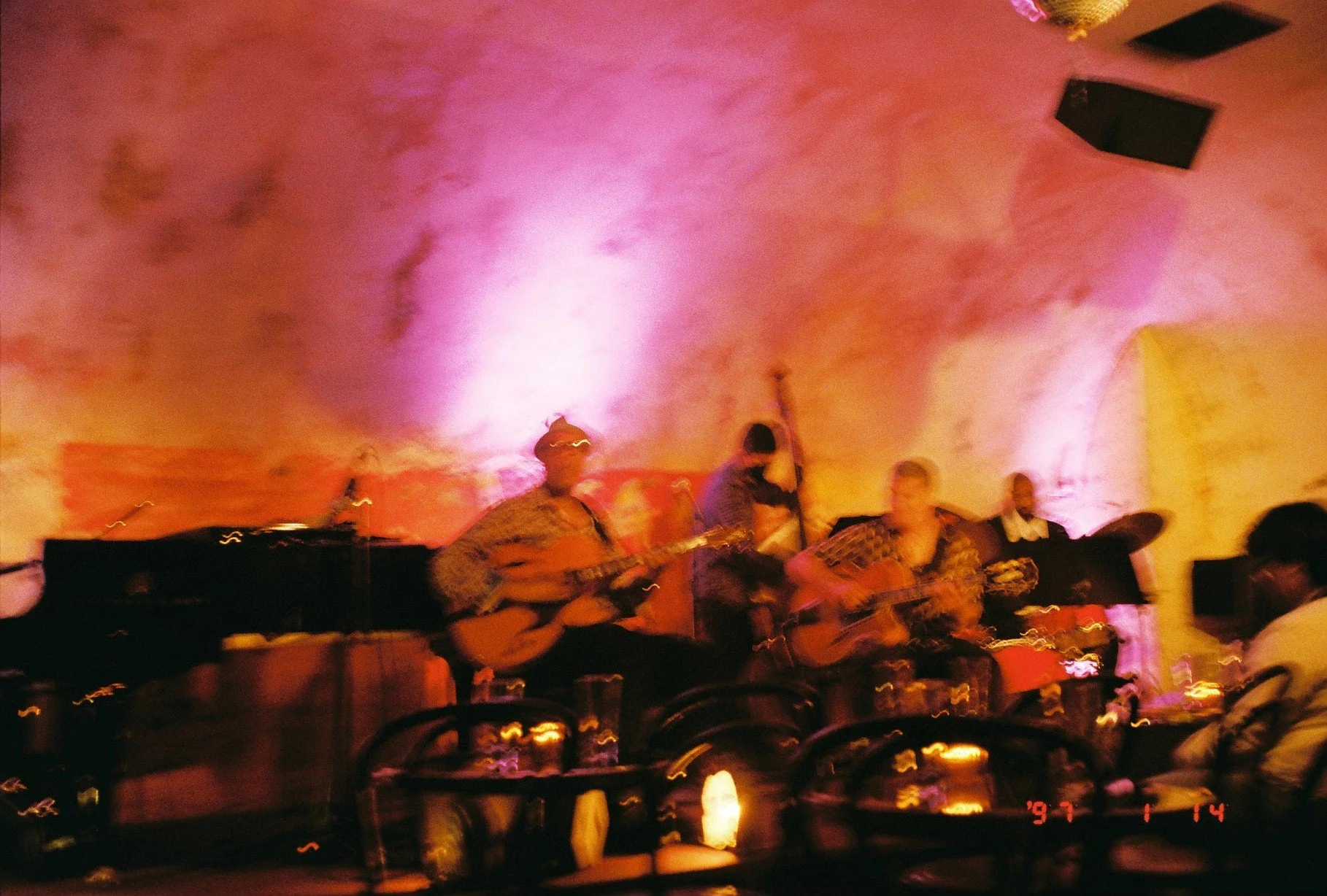 A live musical performance indoors with three musicians playing guitars on a dimly lit stage with pink and purple lighting, and an audience seated nearby.