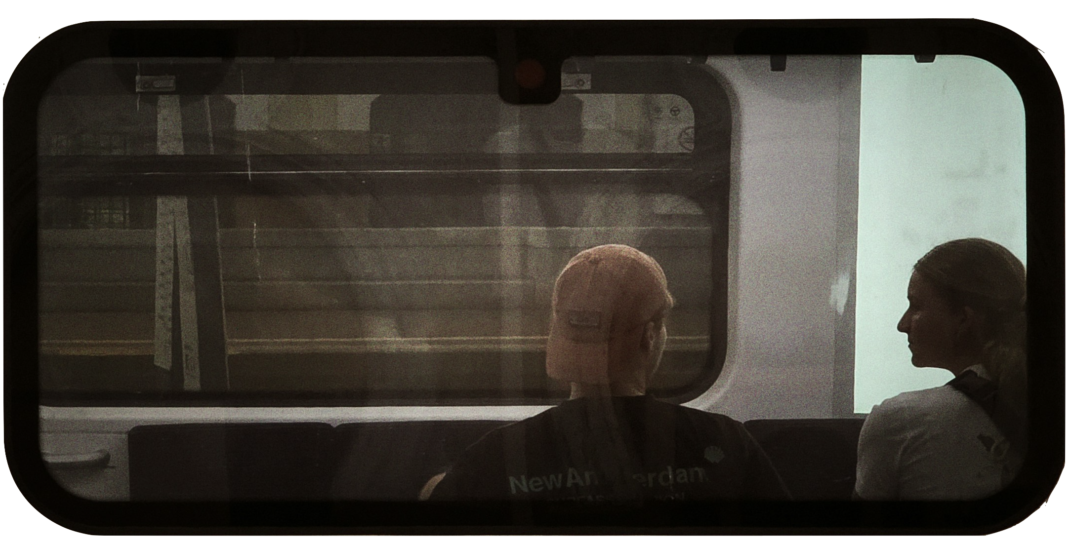 Two women sitting on a train, seen from the back through a window. One has pink hair and the other has long dark hair tied back.
