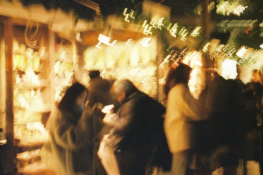 Blurred night scene of people walking and shopping outdoors, illuminated storefronts, and street lights.