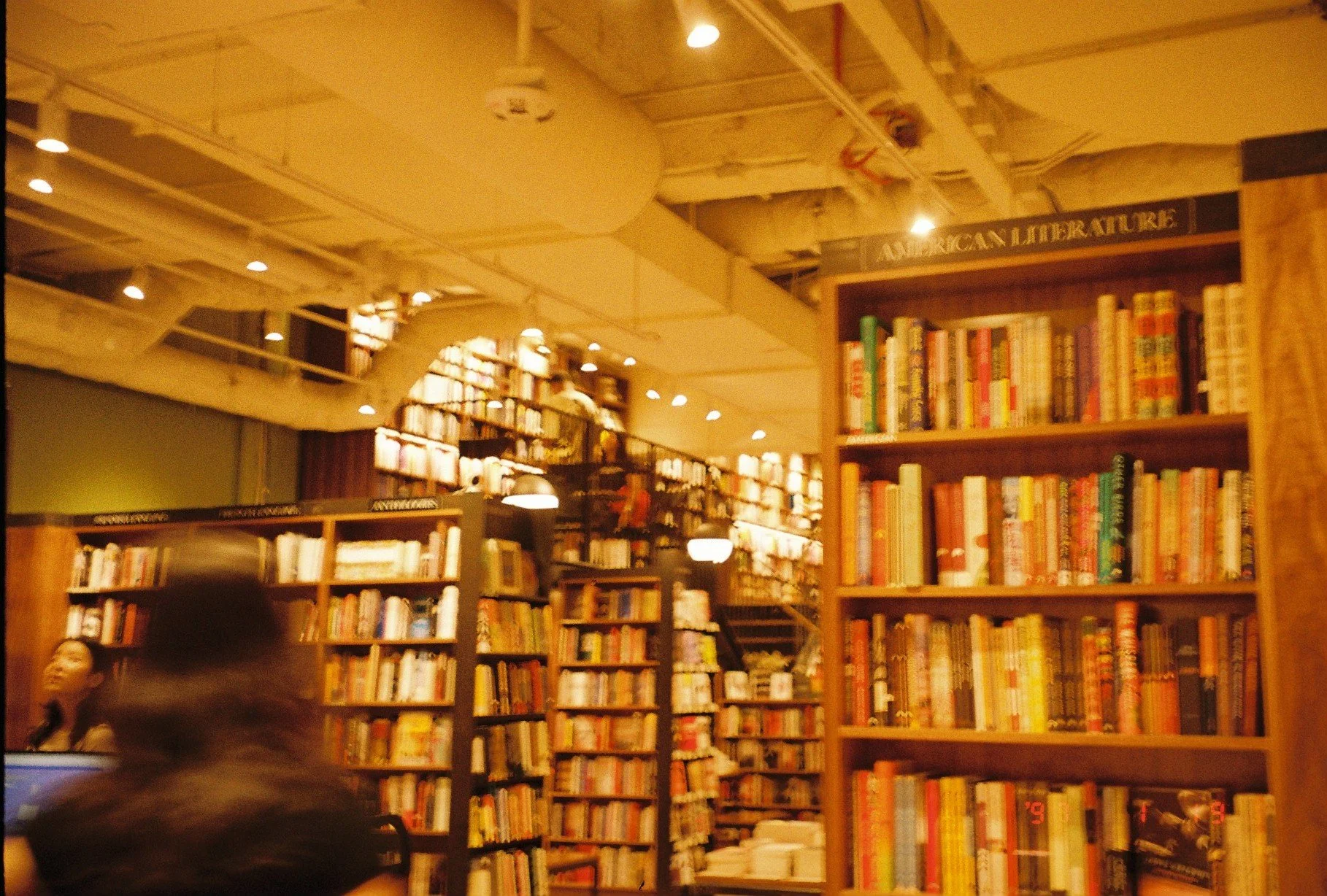 A bookstore with wooden shelves filled with books, labeled 'American Literature,' and people browsing.