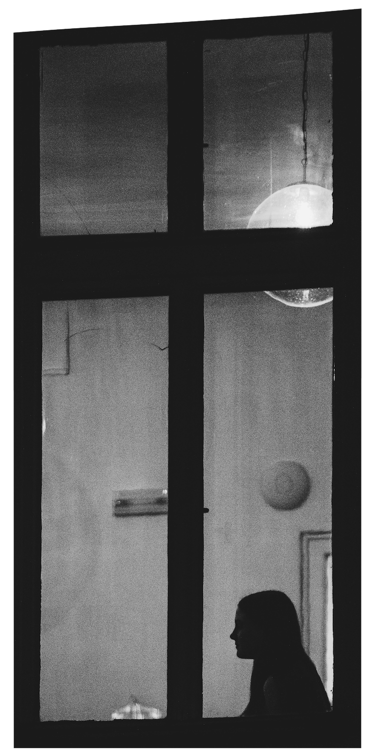 Black and white photo of a woman sitting inside a room, viewed through a window with four panes, showing the silhouette of her profile against the light.