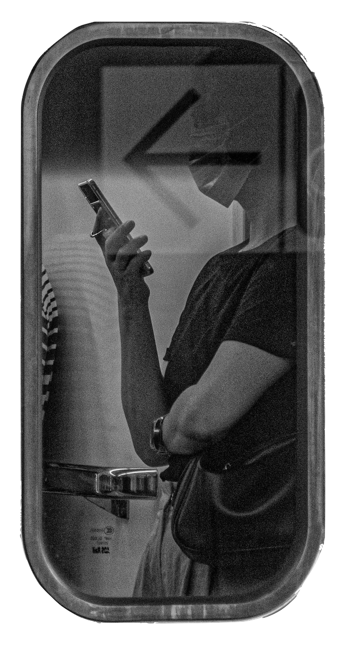 A person standing in front of a mirror, looking at their phone. The reflection shows a large wall clock behind them, with a woman wearing a dark shirt and carrying a handbag.