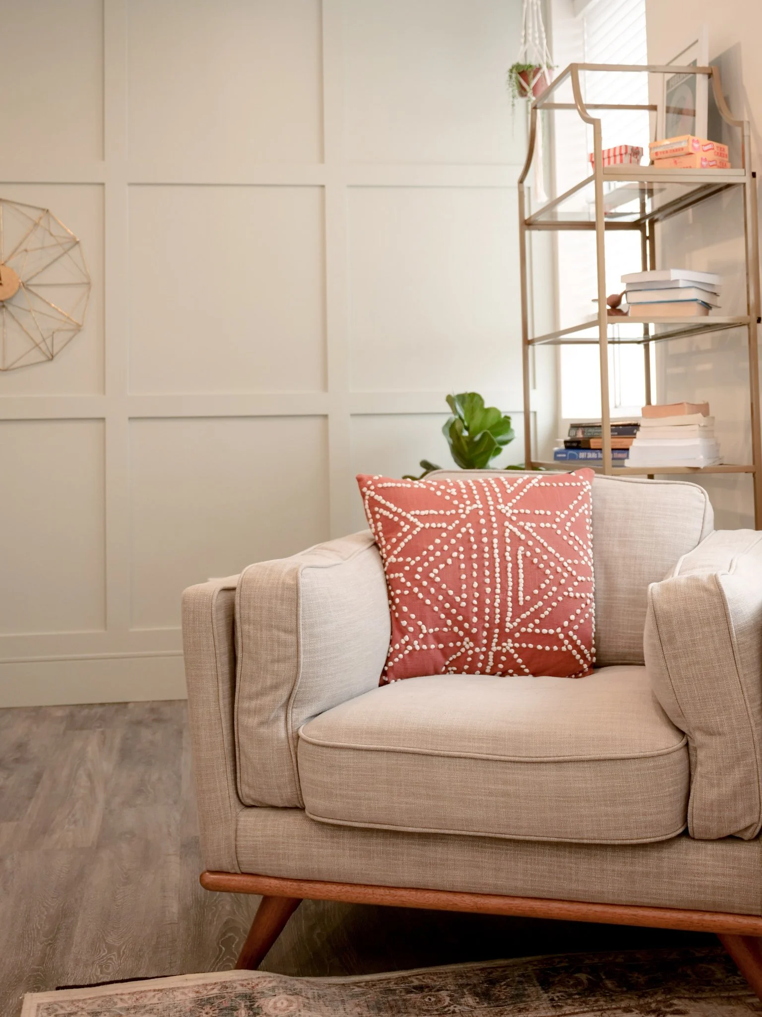 A beige armchair with a pink patterned pillow, a bookcase with books and a potted plant in the background, and a light-colored wall with decorative wall art.