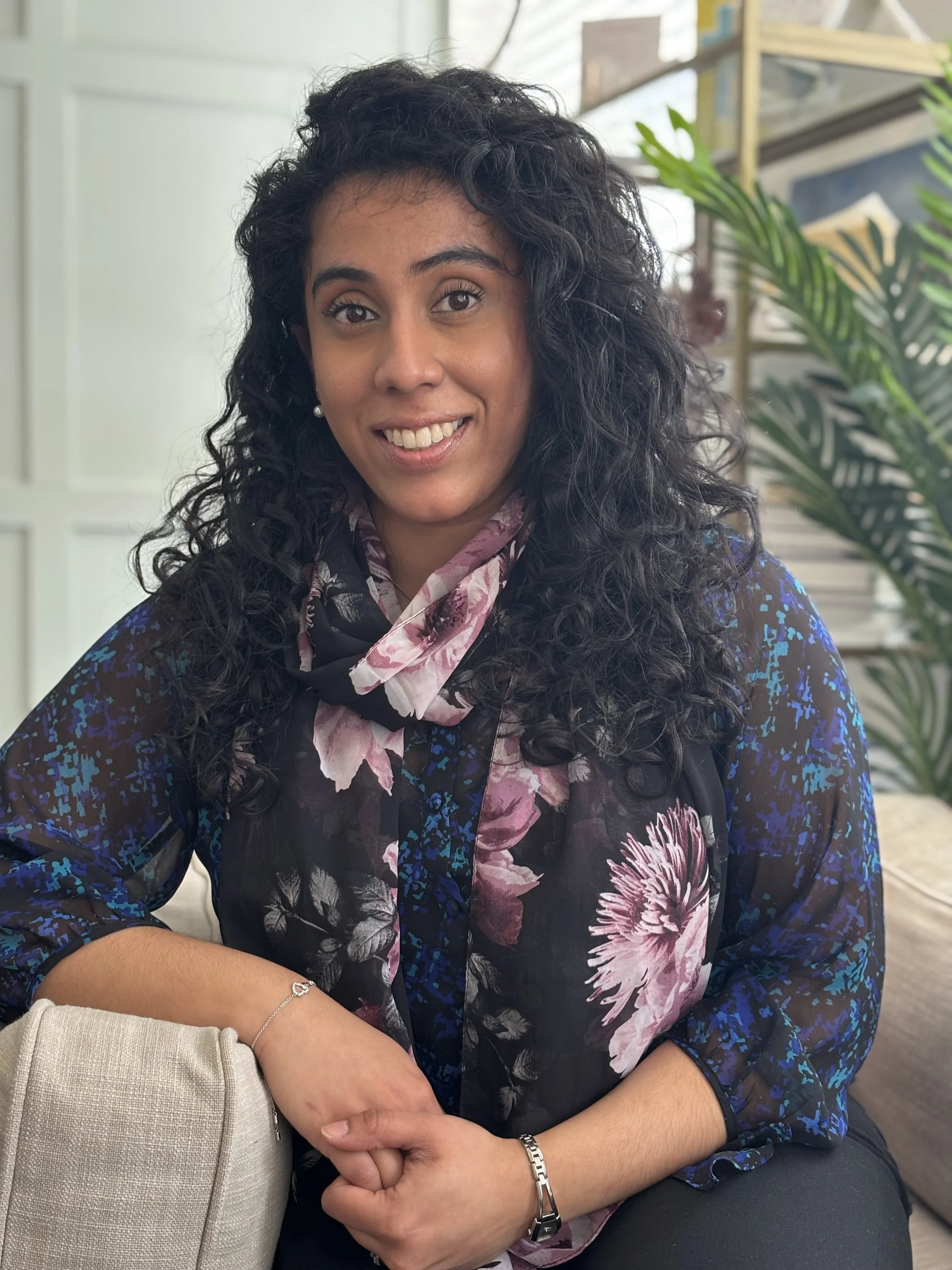 A woman with curly black hair, wearing a floral scarf and a dark blue patterned top, sitting indoors near plants and smiling at the camera.