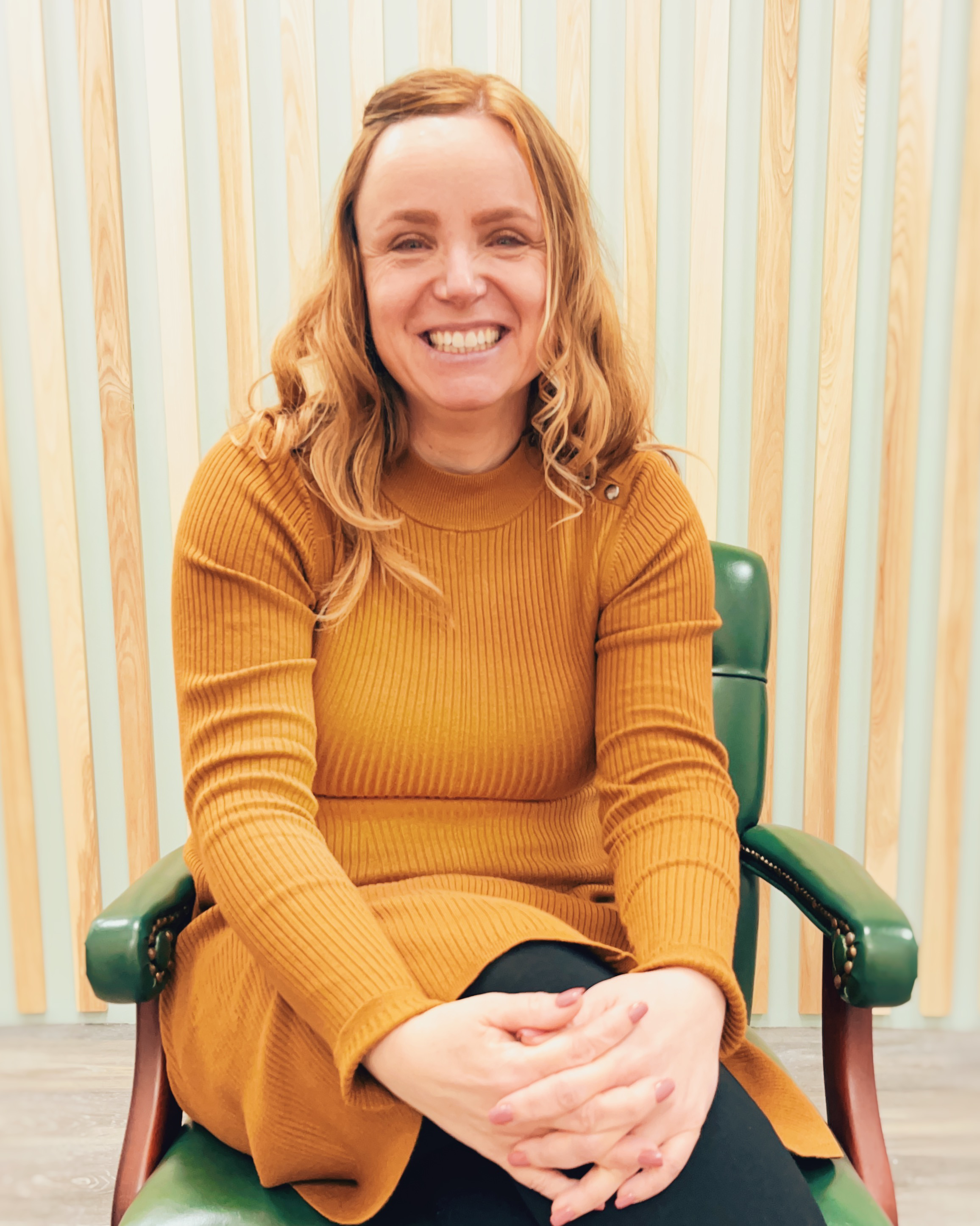 A woman with light skin and red hair, smiling and sitting in a green chair in front of a wooden slat wall.