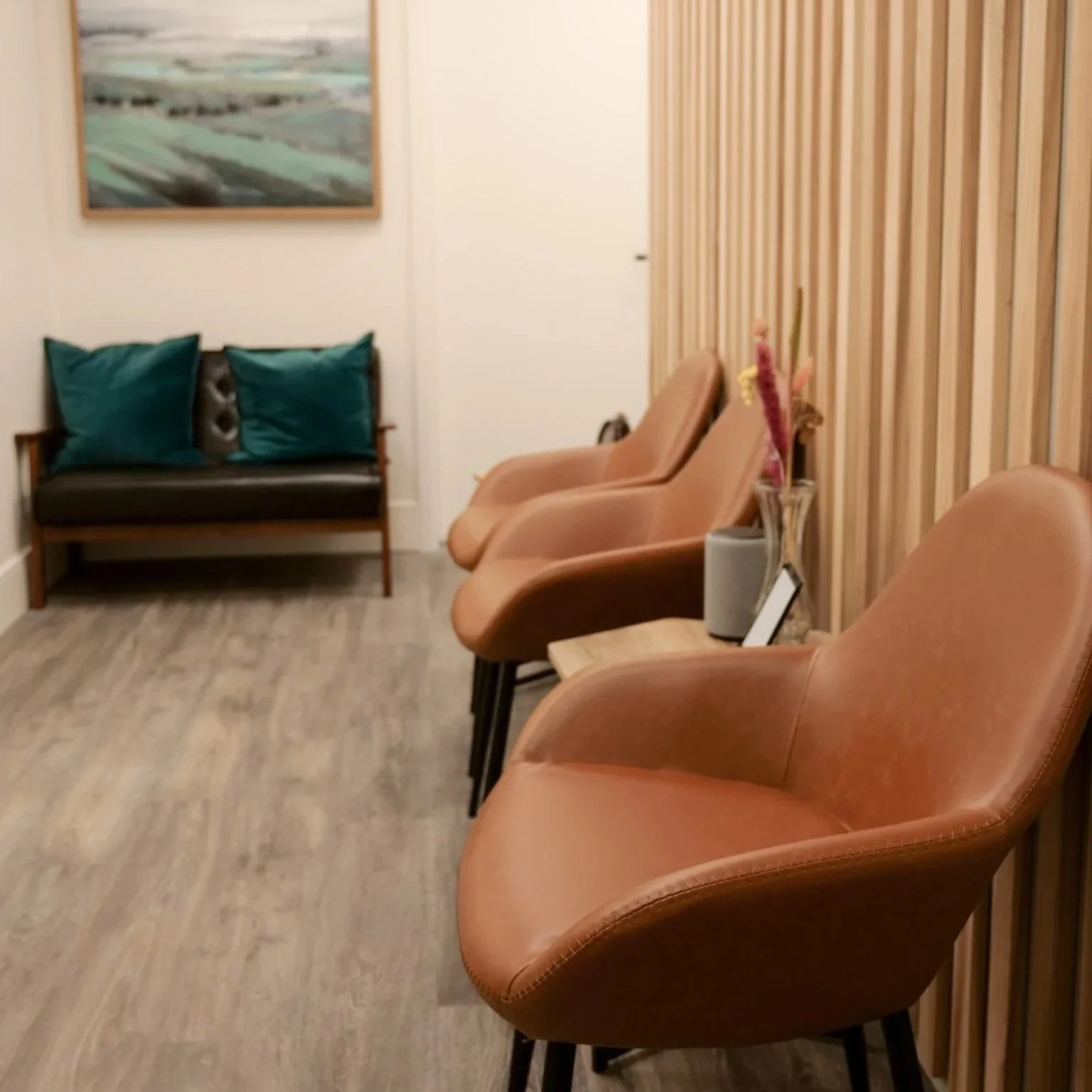 Waiting area with leather chairs, a bench with teal pillows, a flower arrangement, and wall art in a room with wood paneling and hardwood floors.