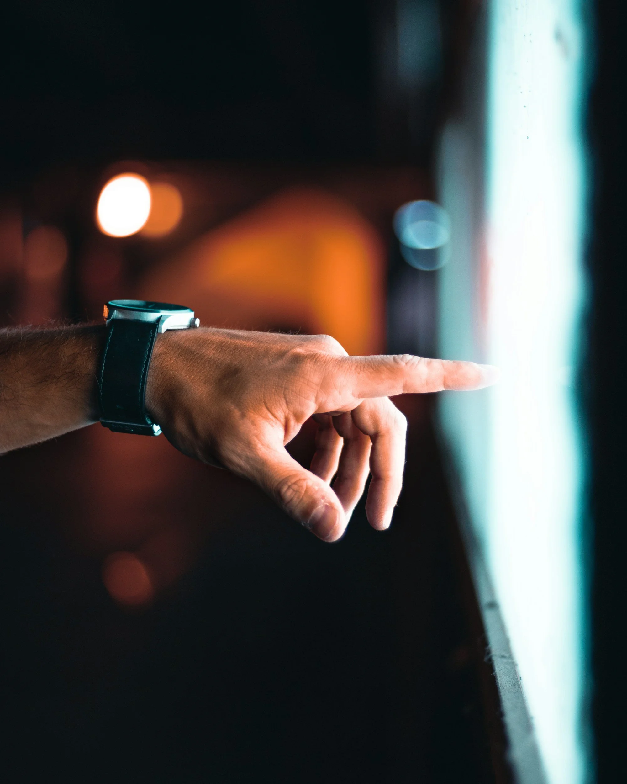 A person wearing a smartwatch is touching or pointing at a bright touchscreen display or monitor in a dimly lit environment.