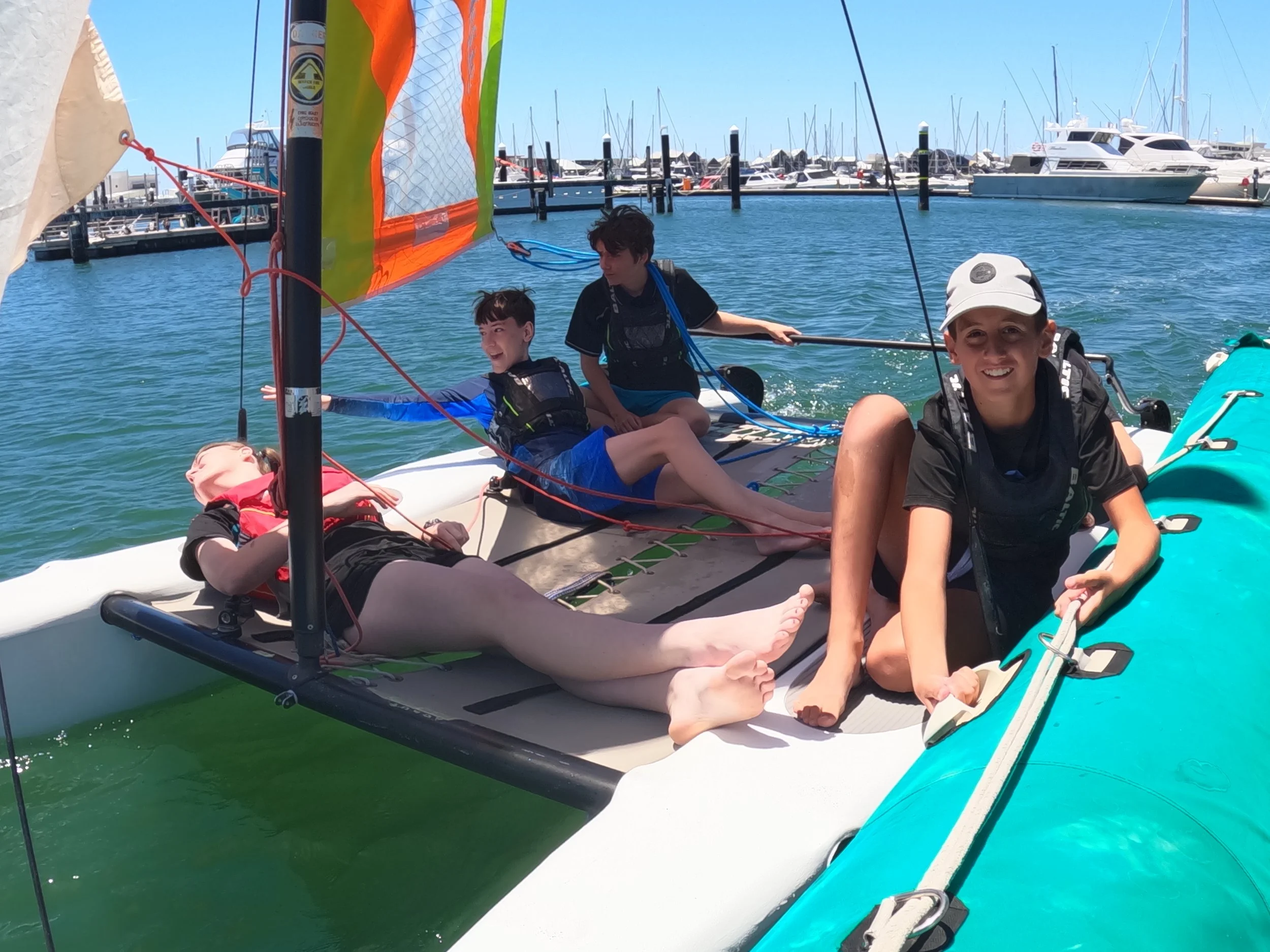 Group of five children on a small sailboat in a marina with several yachts and sailboats in the background under a clear blue sky.