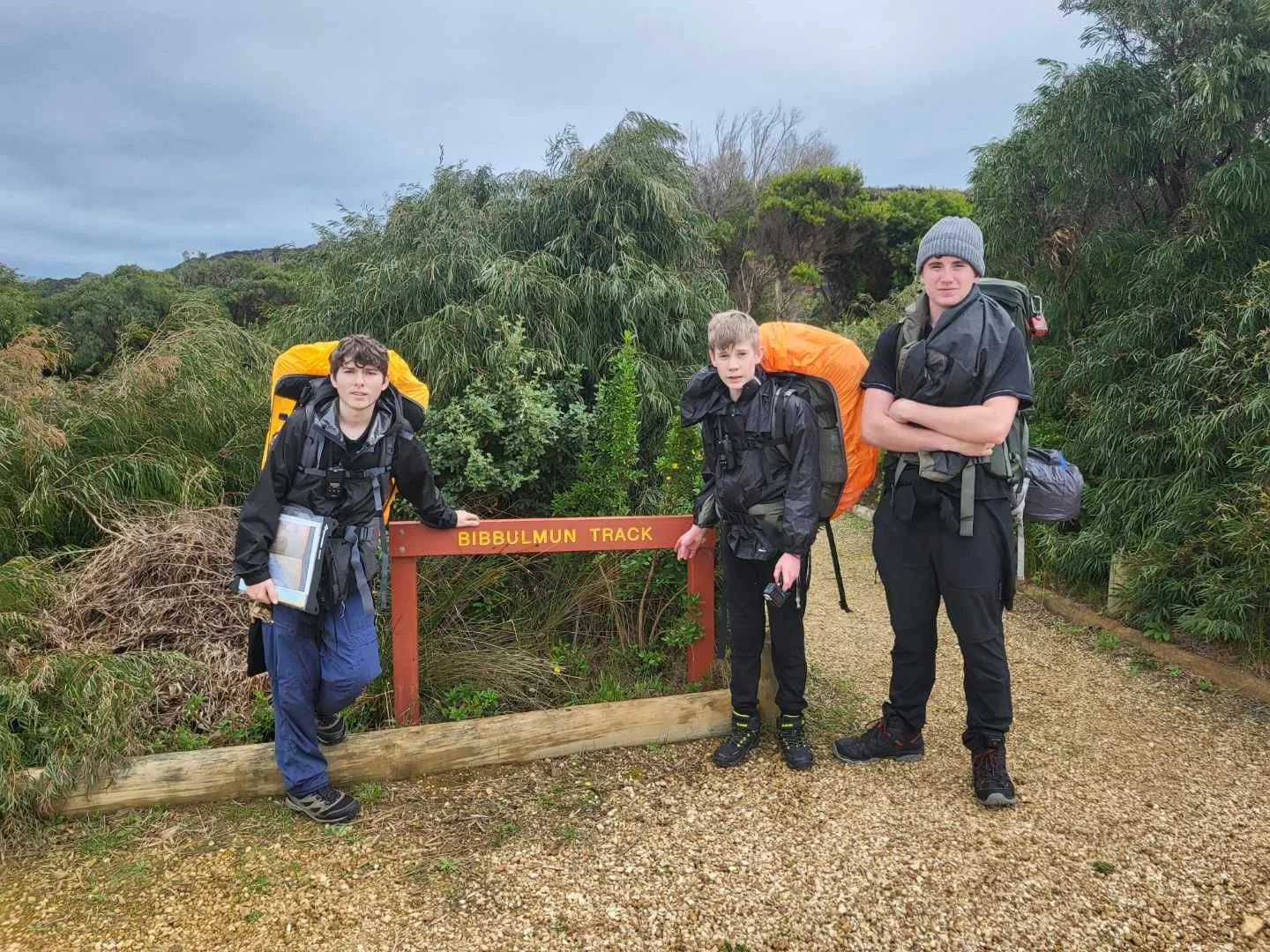 Three young men with backpacks standing on a trail at Bibbulmun Track, surrounded by green bushes and trees, under a cloudy sky.