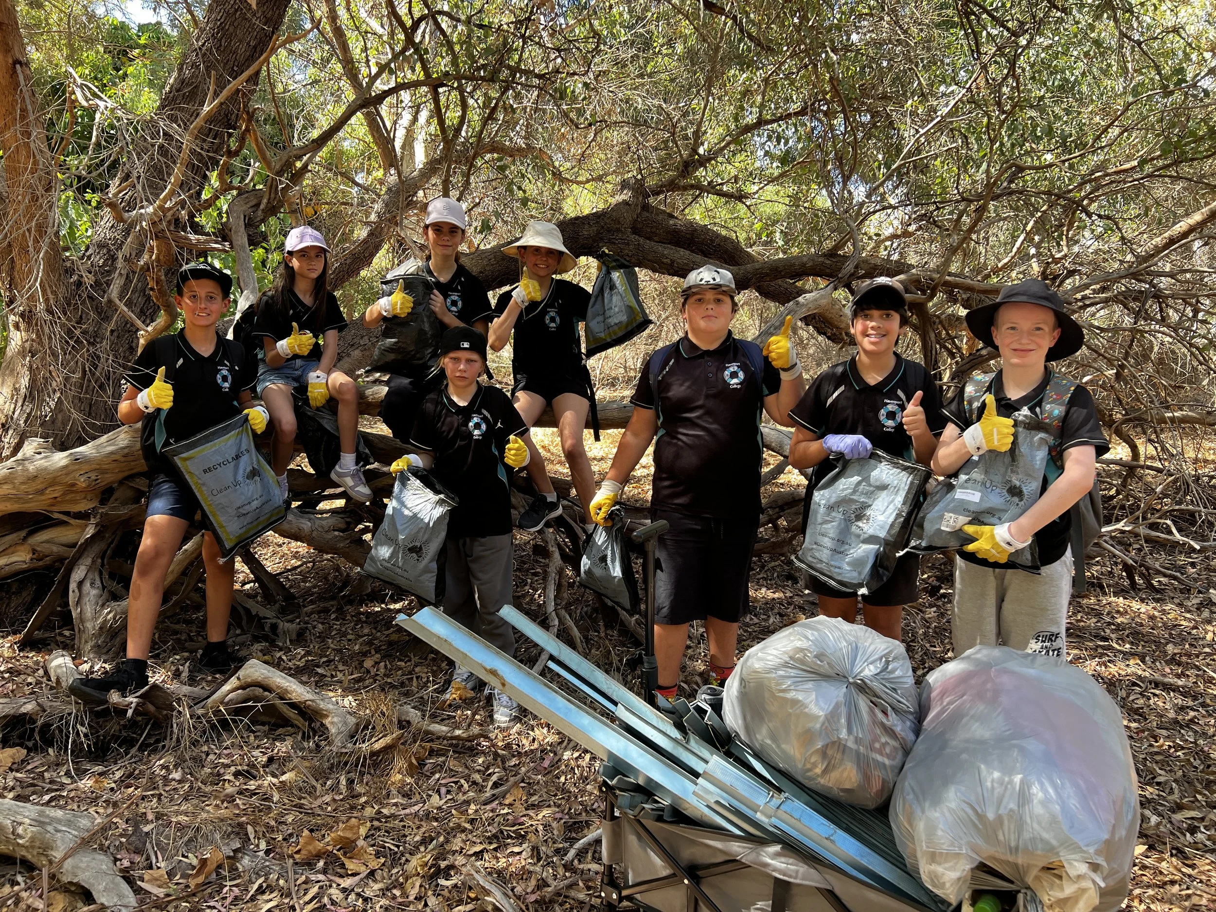A group of ten children in black uniforms and yellow gloves are posing for a photo in a wooded area. They appear to be participating in a cleanup activity, as they are holding trash bags and some are giving thumbs-up gestures. There are two large bags of collected trash and a small cart with more trash bags in front of them.