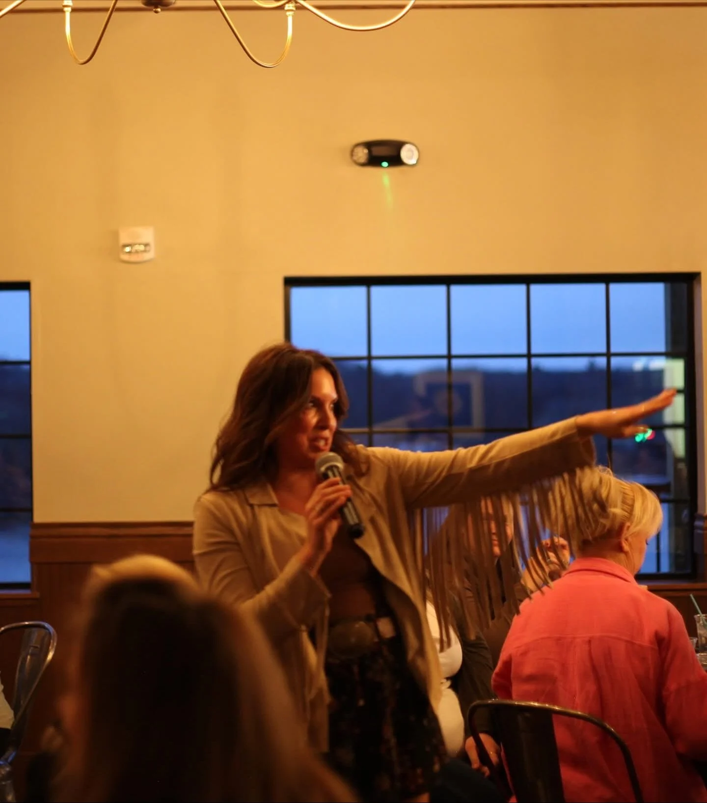A woman with brown hair speaking into a microphone during an indoor event, with her arm extended outward. Several seated people are visible in the foreground, and large windows showing a dusk sky are in the background.