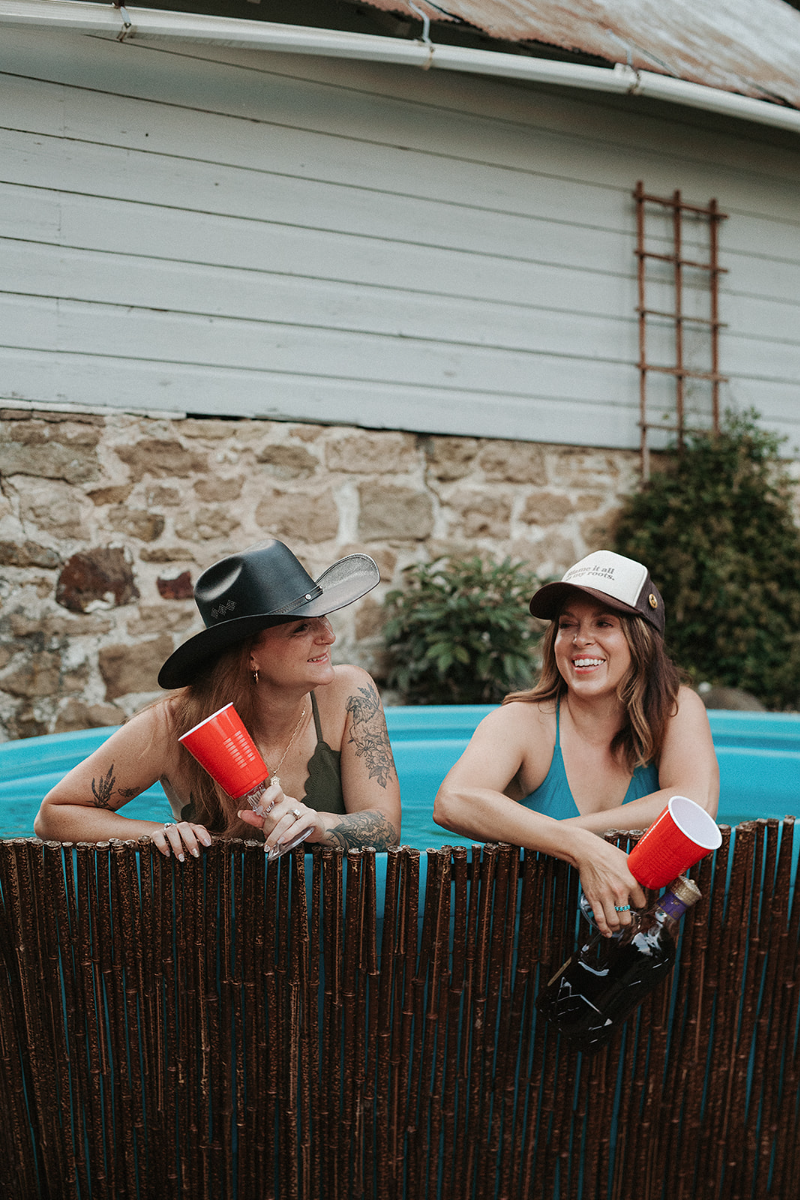 Two women in a small above-ground pool outdoors, smiling and chatting, with drinks in red cups. One woman wears a black hat and a dark swimsuit; the other wears a cap and a blue swimsuit.
