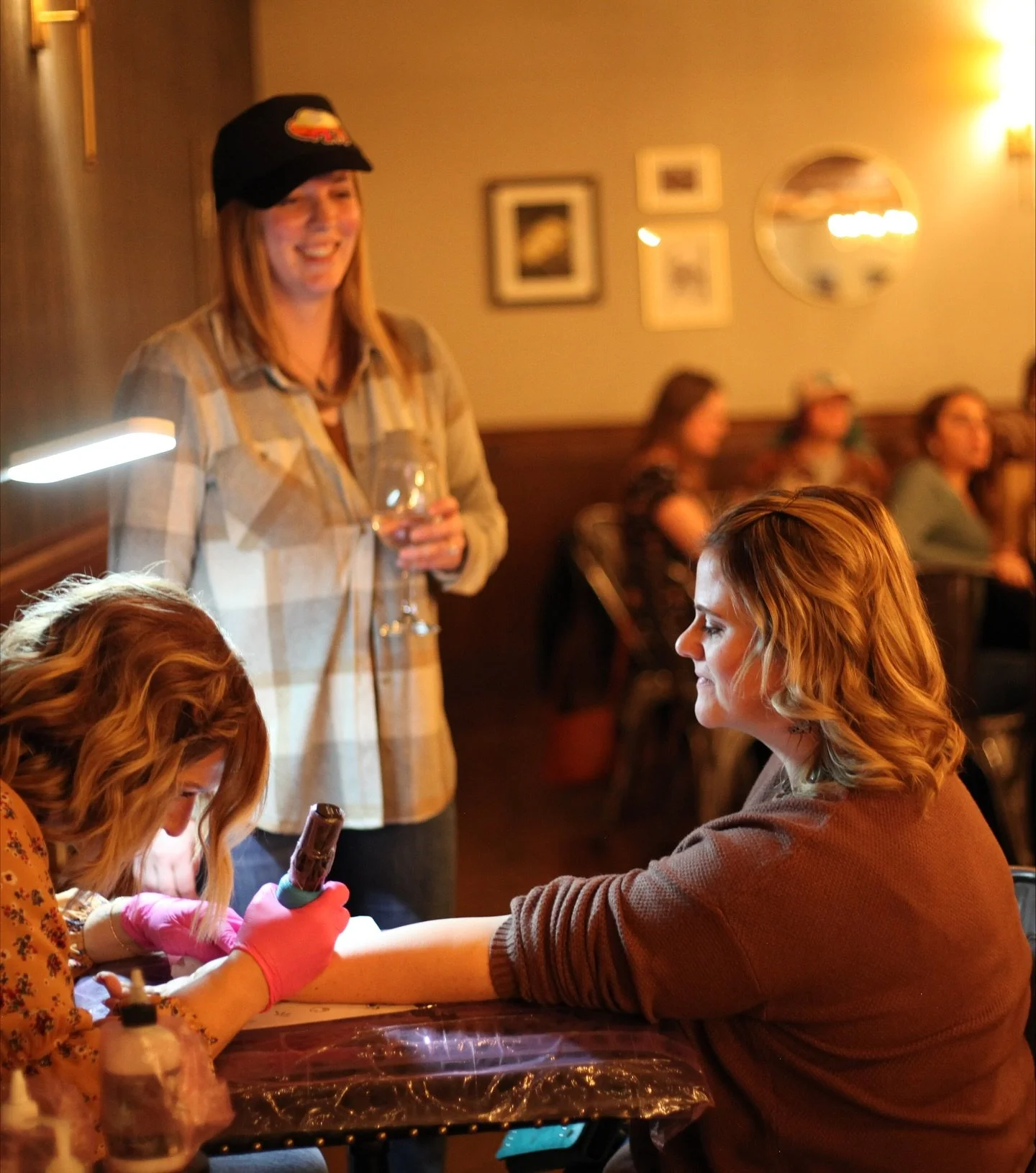 A woman getting a tattoo on her arm at a tattoo parlor, with several people sitting in the background.