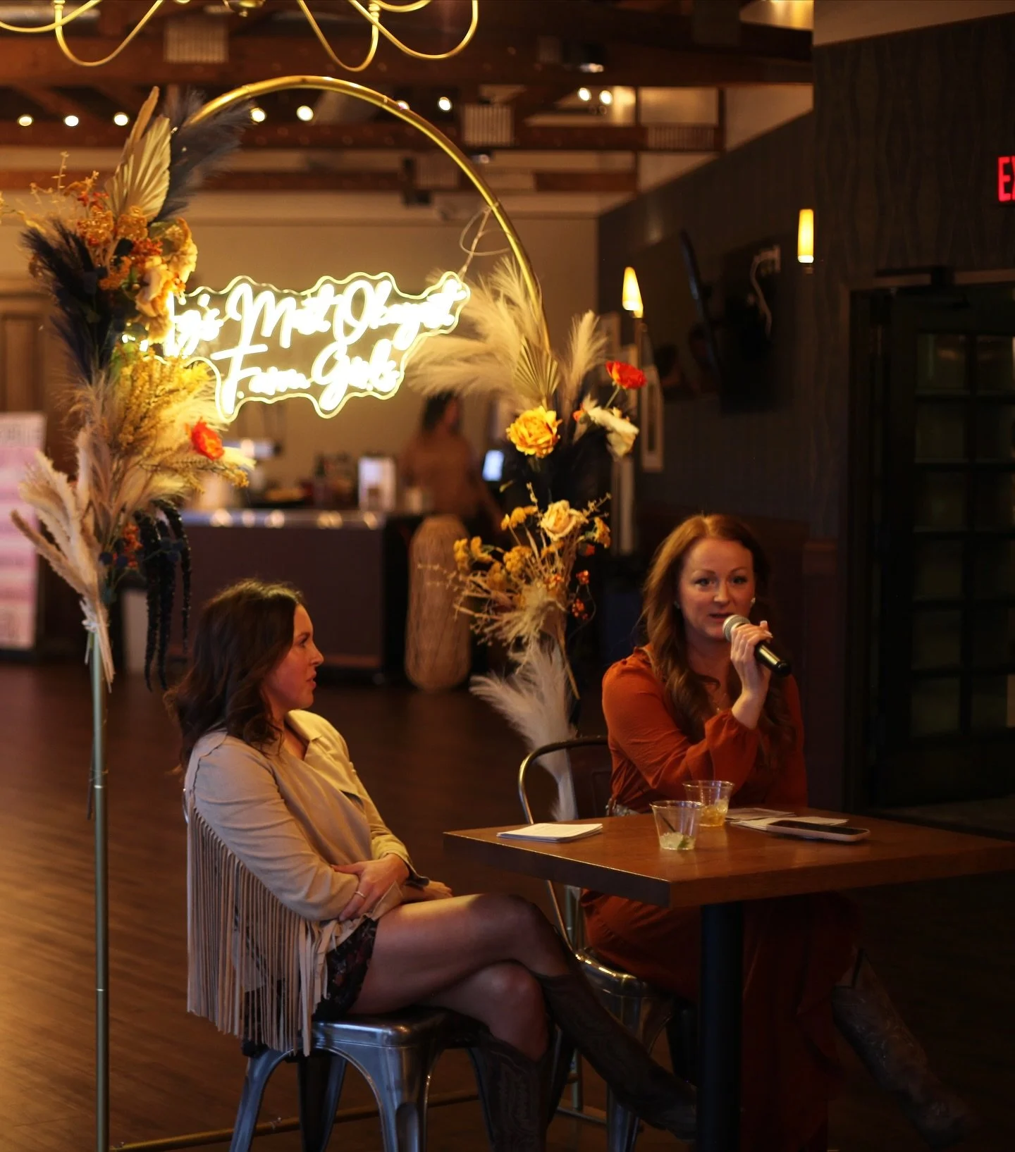 Two women sit at a table inside a dimly lit venue, with one woman speaking into a microphone. There are floral arrangements and a neon sign that reads 'The Moth Dinner, From Gilt' in the background.