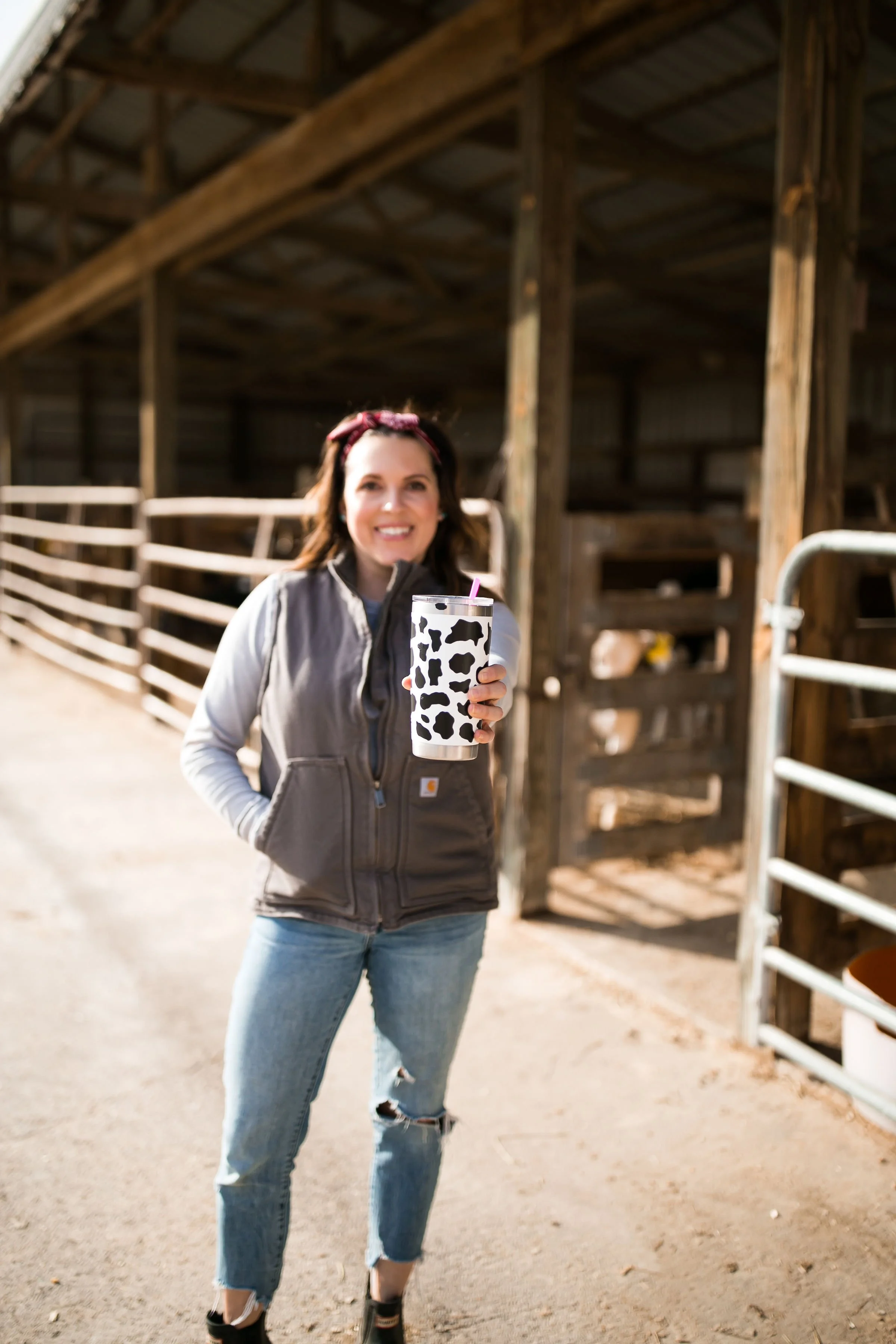 Woman standing inside a barn holding a cow-print drink tumbler and smiling.