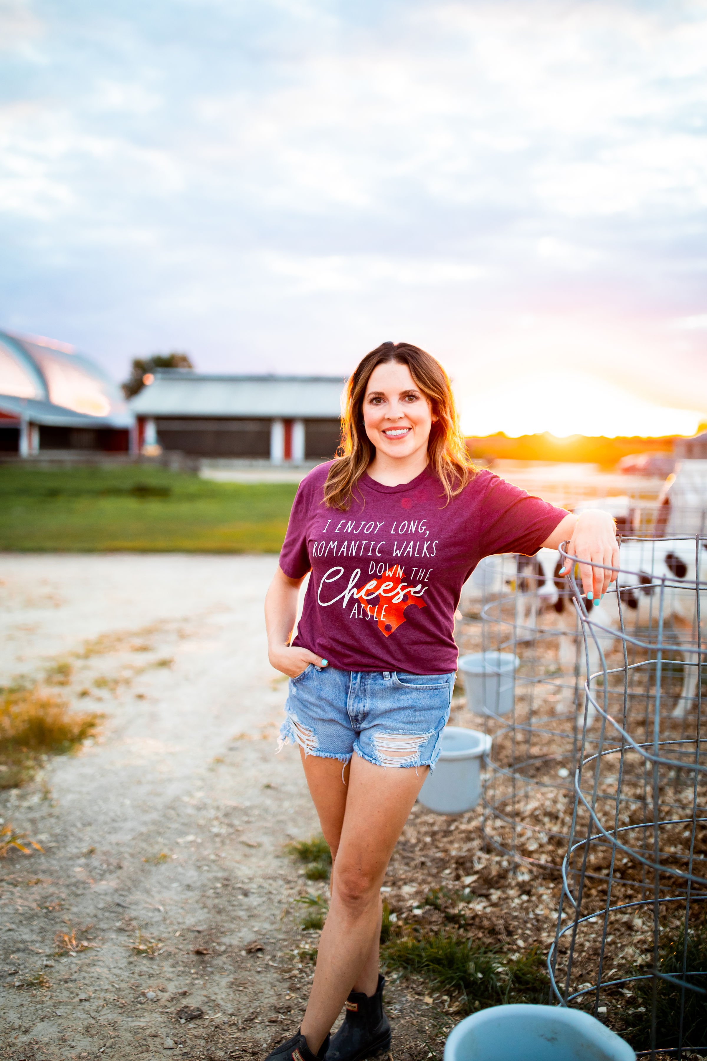 A woman standing outdoors near a wire fence at sunset, wearing a maroon t-shirt with text and ripped denim shorts, smiling at the camera.