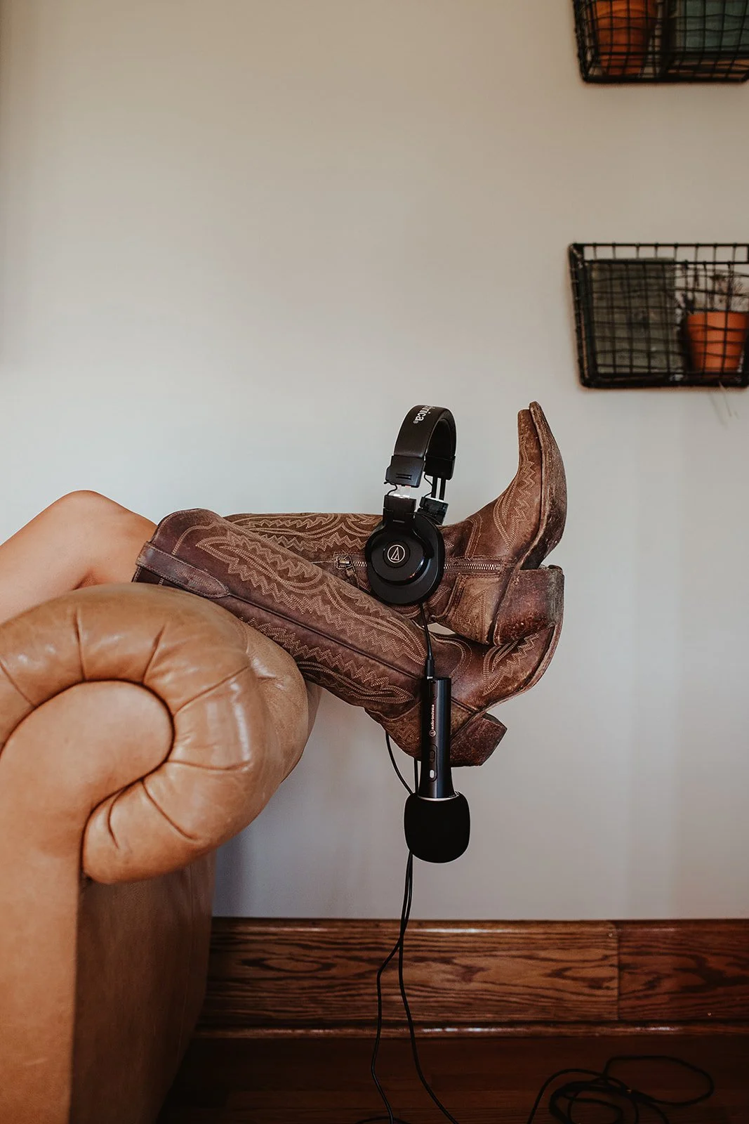 A pair of brown cowboy boots resting on the armrest of a tan leather sofa, with a black pair of headphones hanging on the boots. The background shows a plain wall with two black wire wall shelves, one holding a small potted plant.