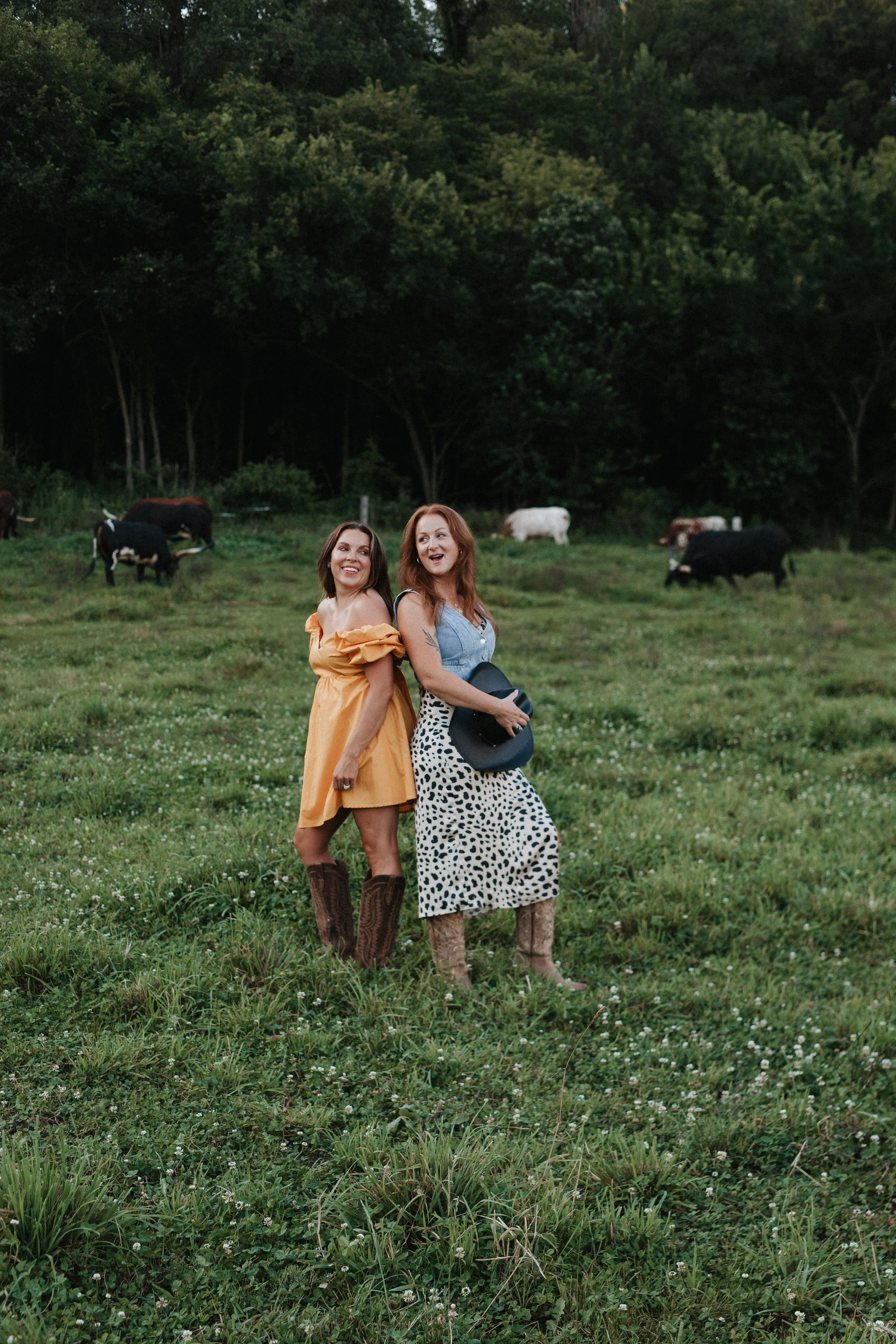 Two women standing in a grassy field with cows grazing in the background, smiling and enjoying the outdoors.