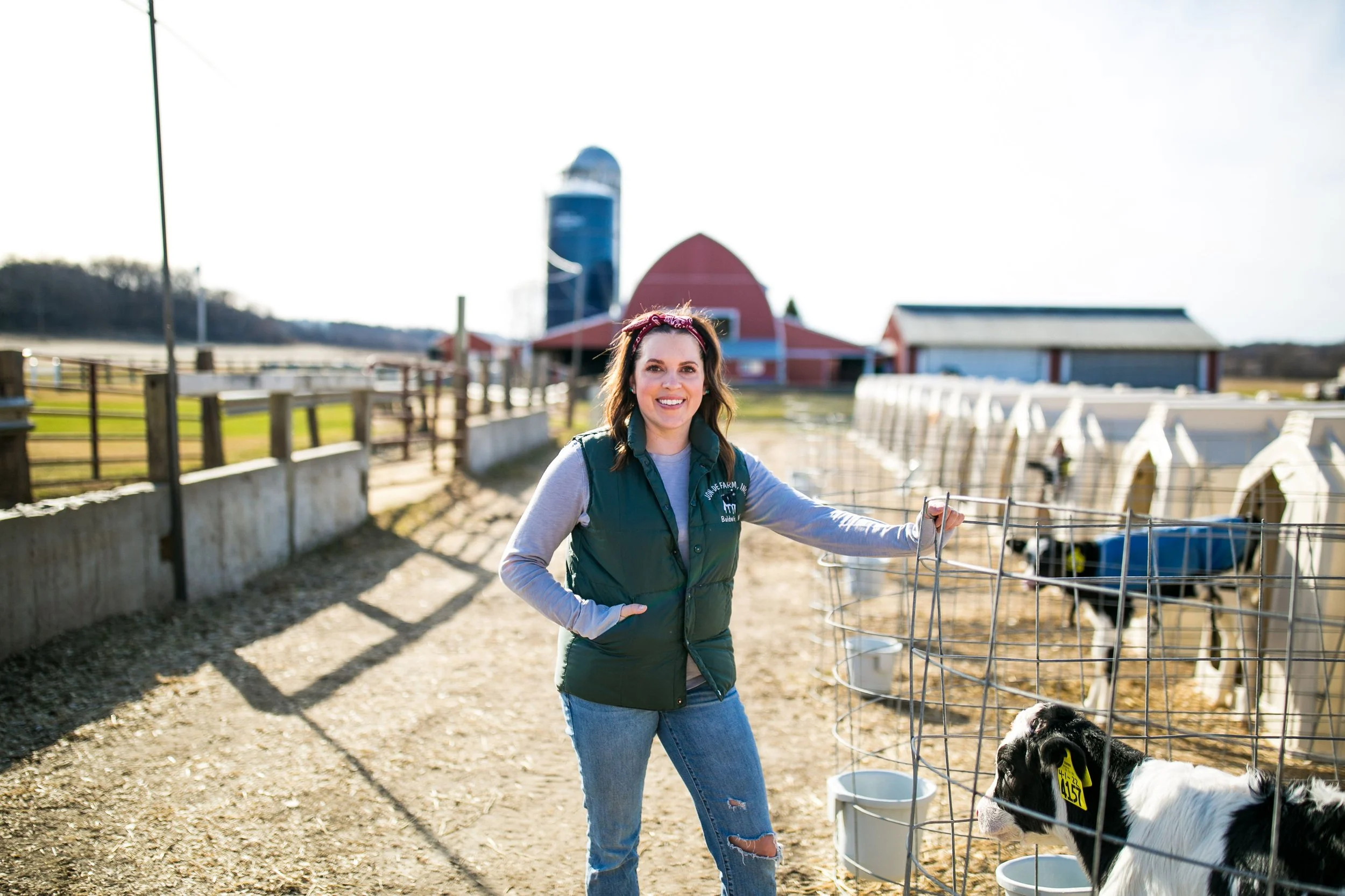 A woman smiling on a farm with black and white calves in front of her, some calves in a wire enclosure, with barn and farm buildings in the background.