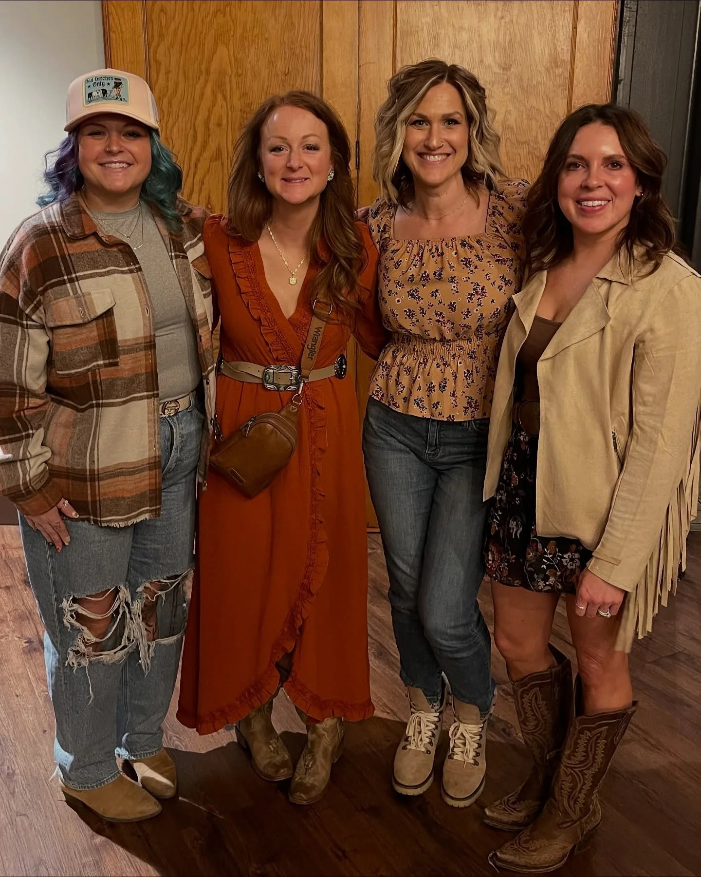 Four women standing indoors, smiling at the camera, wearing casual and country-style clothing.