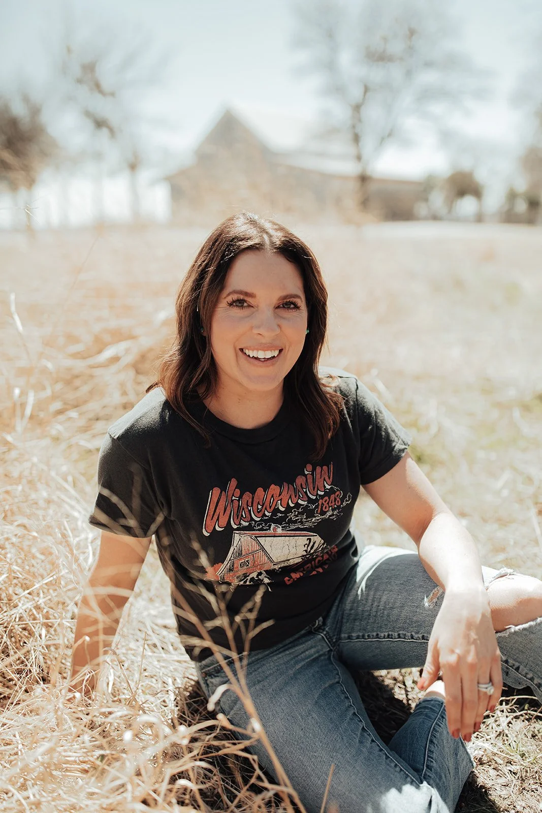 A woman with brown hair sitting on dry grass, smiling at the camera, wearing a black Wisconsin T-shirt and jeans, with a blurred barn and trees in the background.