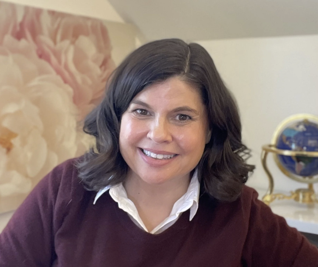 A woman with shoulder-length dark hair, wearing a white collared shirt and a burgundy sweater, smiling at the camera. In the background, there is an abstract floral painting and a decorative globe on a table.
