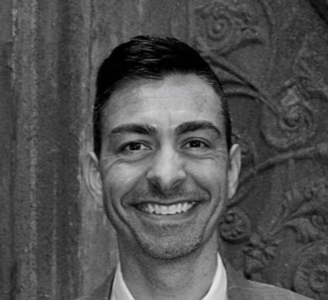 A smiling man with short dark hair, wearing a suit, in front of a decorative carved stone background