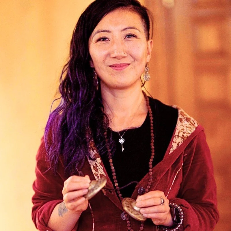 A woman with long, wavy purple-dyed hair and light skin, smiling while holding a crystal ball in each hand, wearing a maroon hoodie with an embroidered design, earrings, a key necklace, and multiple bracelets, standing against a warm indoor background.
