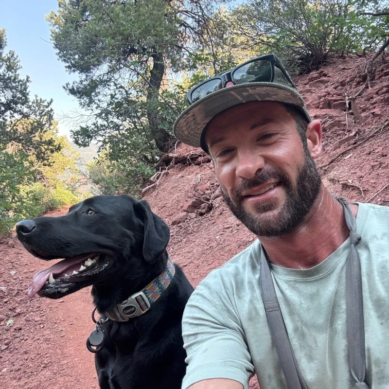A man in a gray t-shirt and cap taking a selfie with a black dog on a dirt trail in a wooded area.