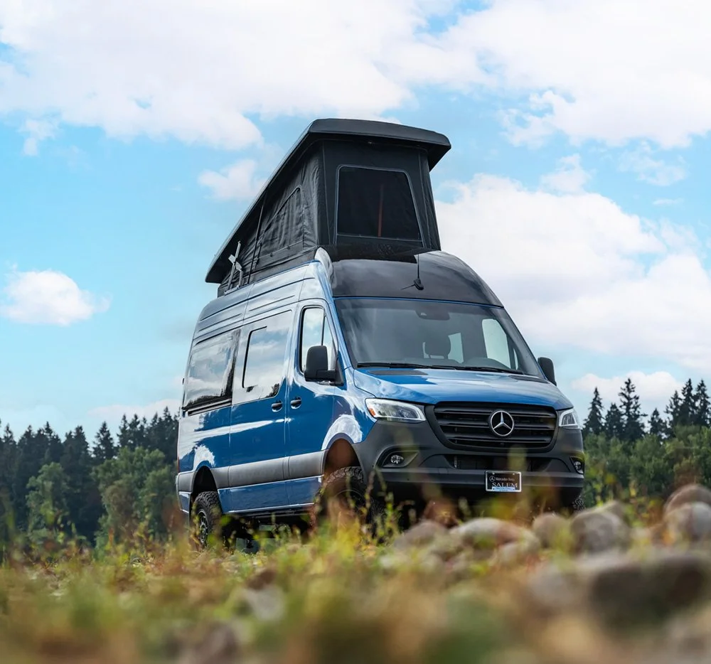 A blue Mercedes-Benz van with a rooftop tent parked in a natural outdoor setting with trees and a cloudy sky.