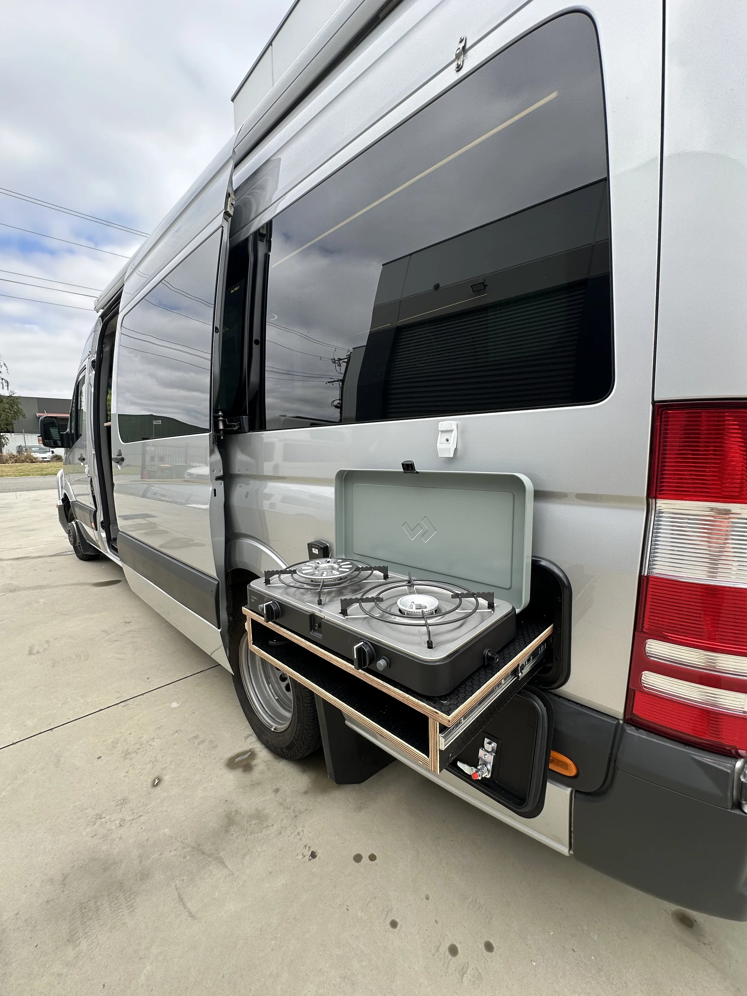 A camper van with an open side door, showing a portable stove attached to the exterior of the van.