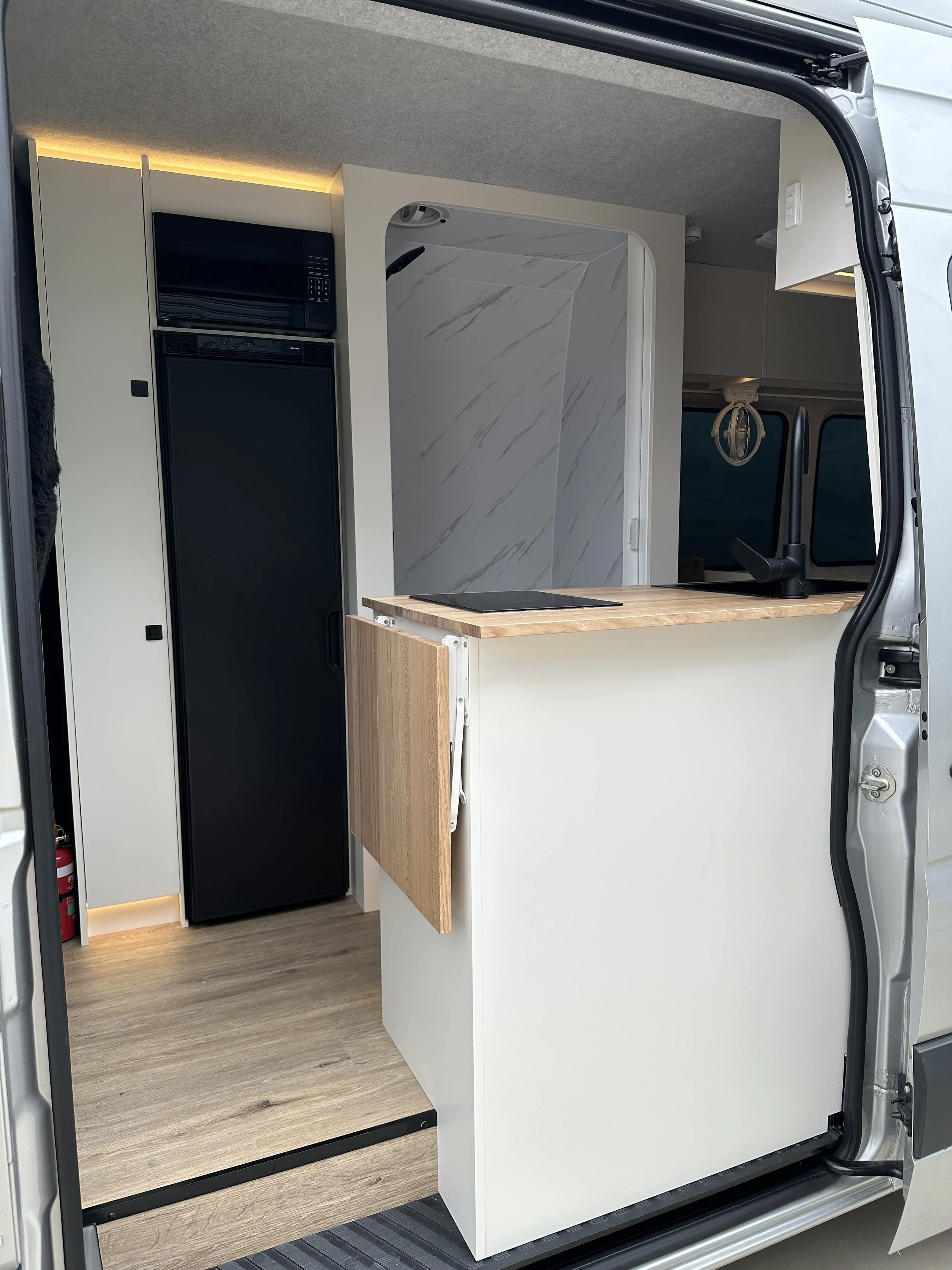 Interior of a compact camper van with a small kitchen area, black refrigerator, microwave, and a wood countertop, with a partial view of a marble-patterned wall.