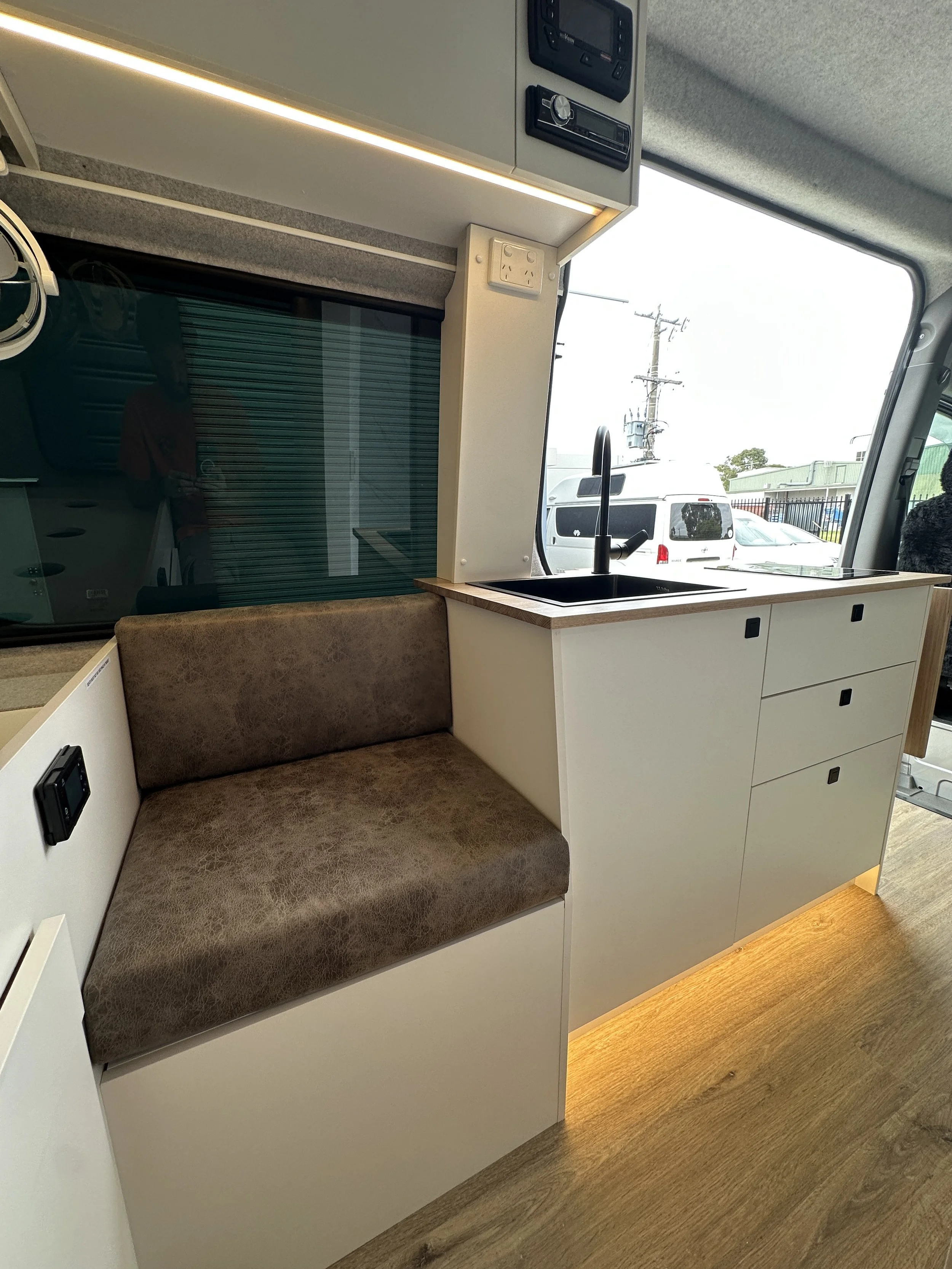 Interior of a small camper van showing a seating area with a brown upholstered bench, a kitchenette with a black sink and modern black faucet, white cabinets with black handles, and a large window with a view of parked vehicles outside.