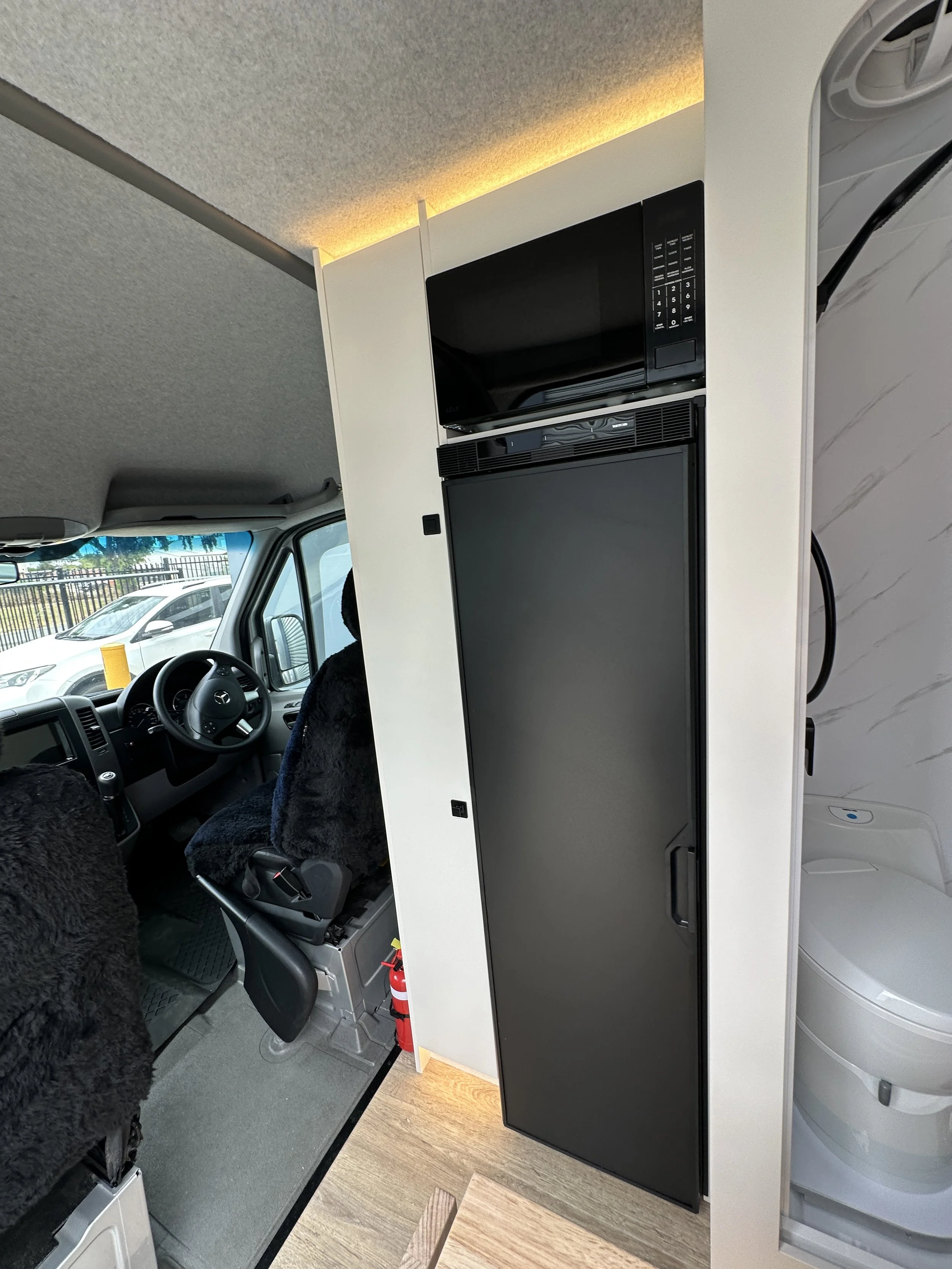 Inside a camper van or RV with a microwave and fridge unit, with cluttered driver’s seat and dashboard, and a partial view of the bathroom area.
