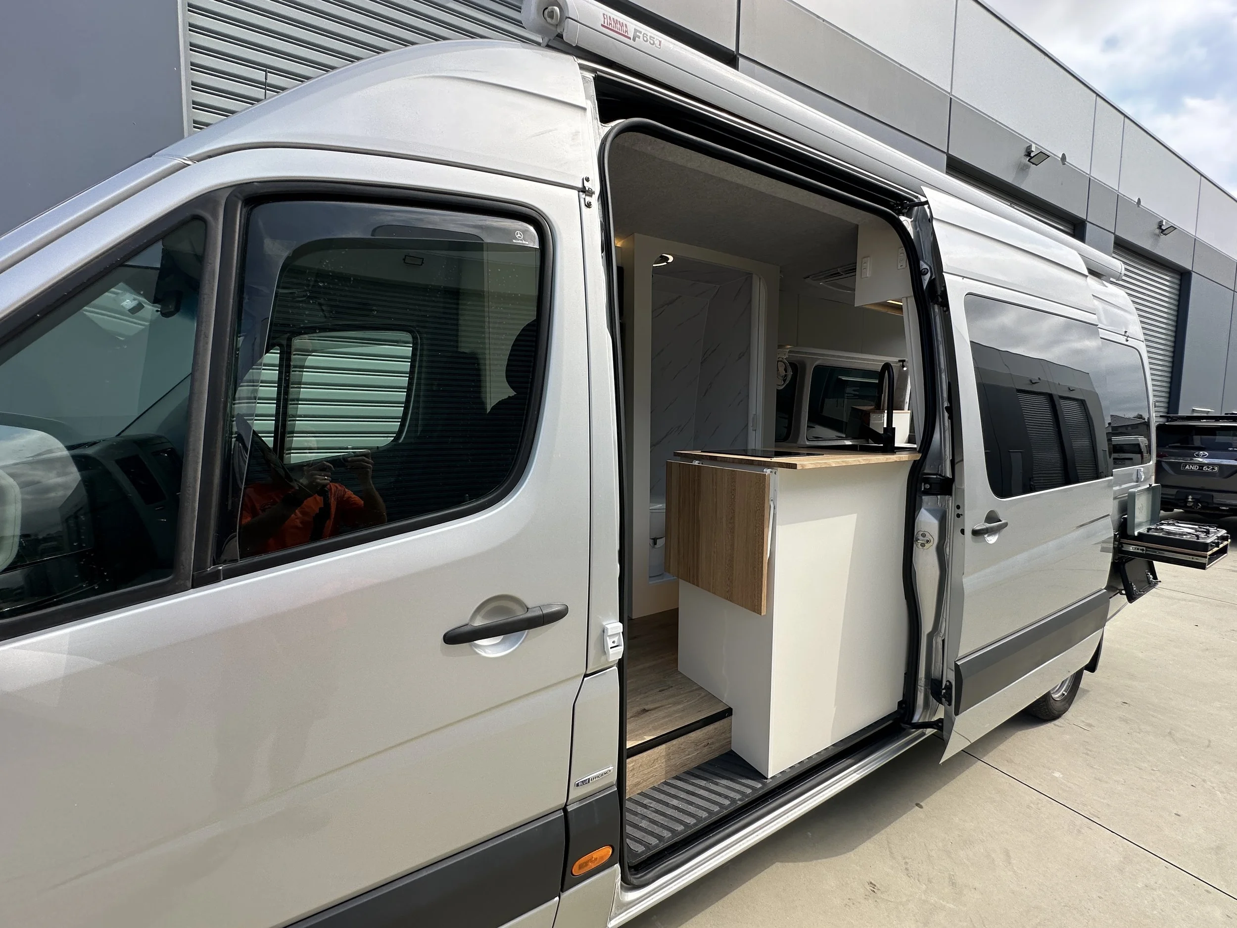 Side view of a silver van with a partially opened sliding door, revealing a wooden kitchen counter and small appliances inside.