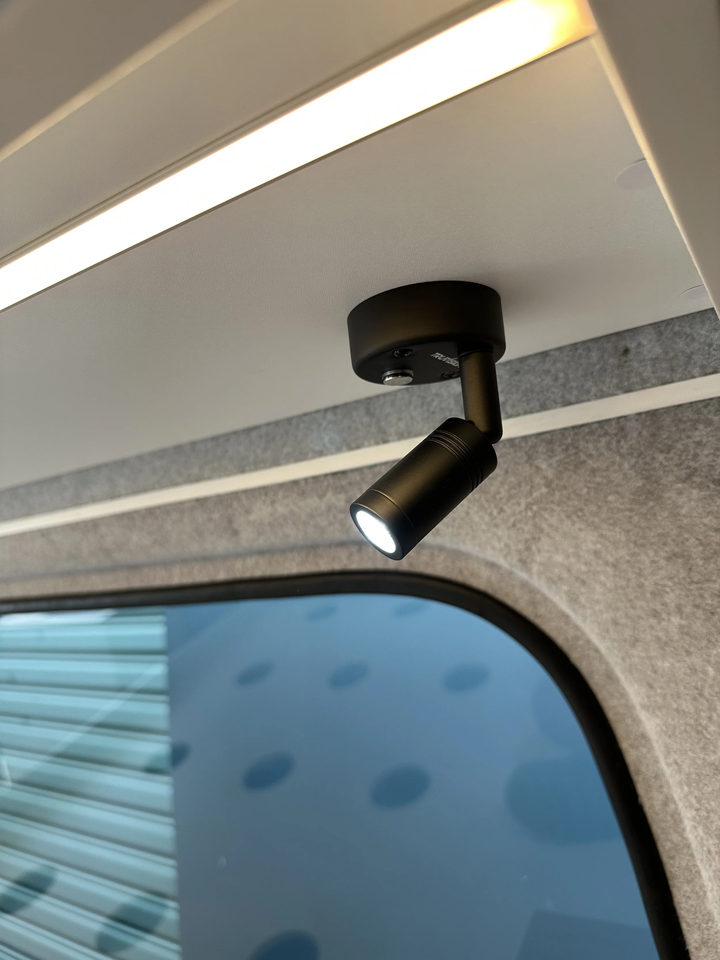 Close-up of a small black spotlight interior ceiling light, mounted on a beige ceiling next to a window with a blue sky and clouds visible outside.