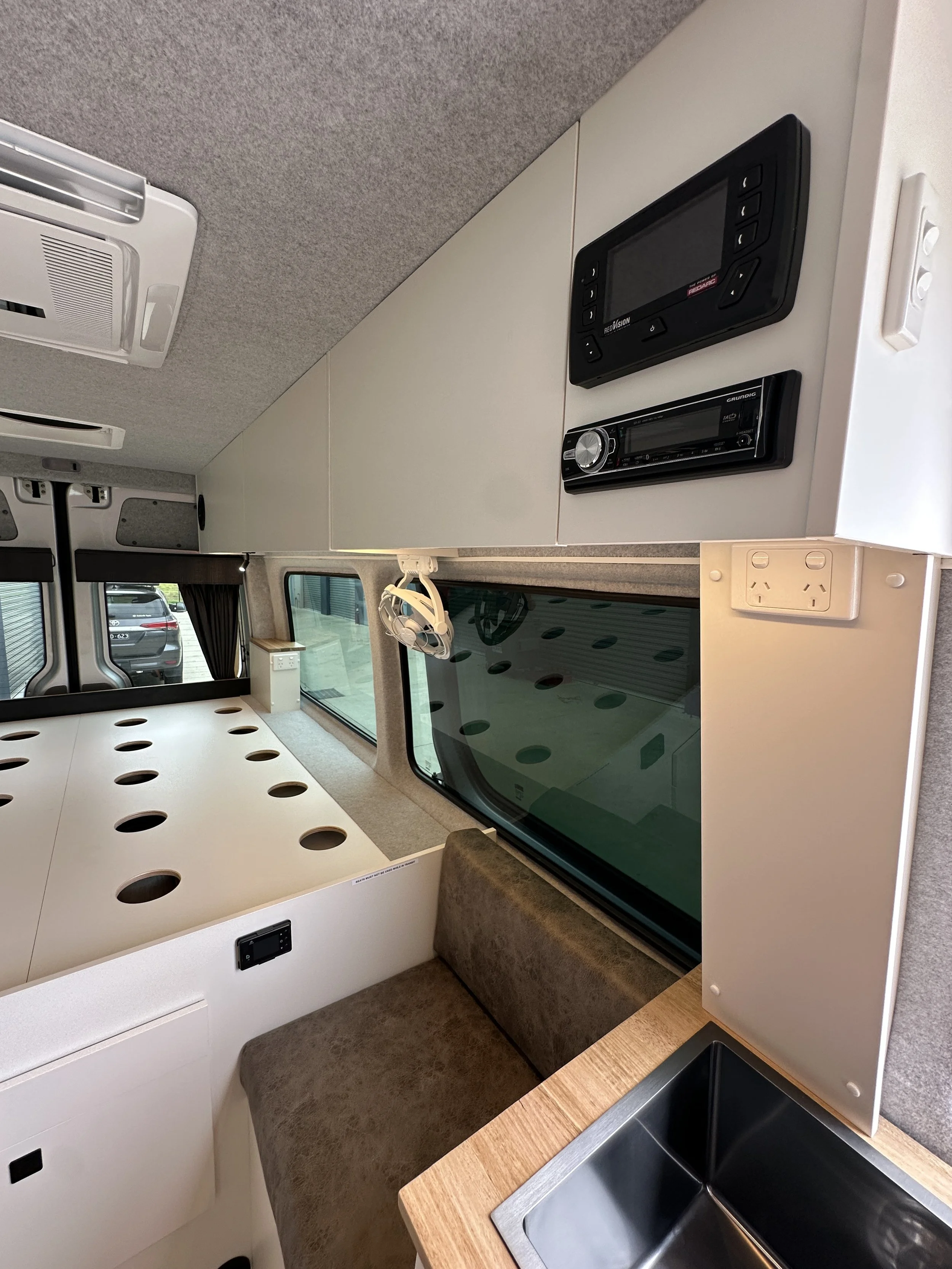 Interior of a campervan or RV with a table with circular holes, a window with curtains, a small sink, a padded seat, and overhead cabinets with electronic devices mounted.