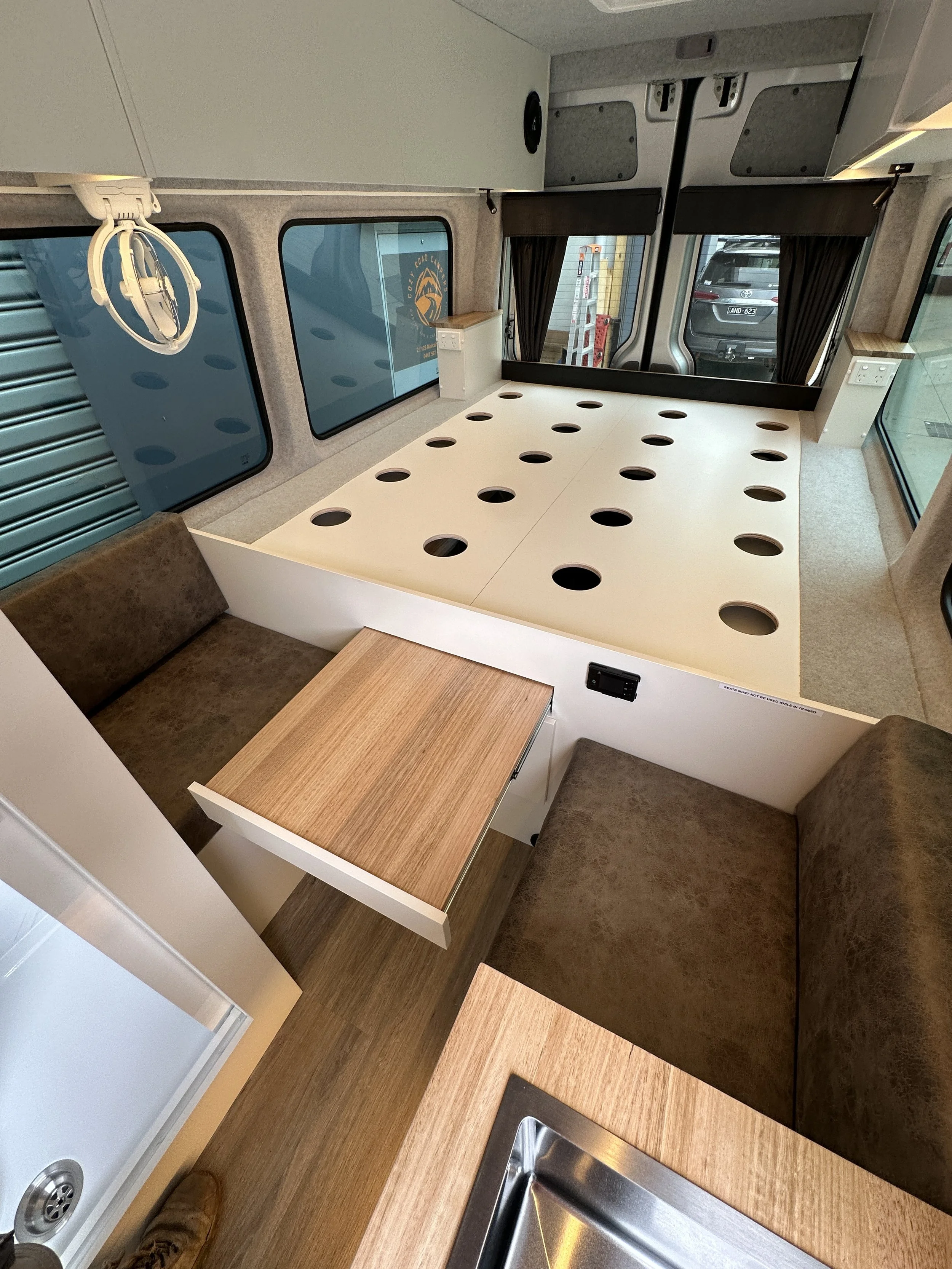 Interior of a camper van with a custom-built fold-out bed with circular cutouts, small dining area with a wooden table and brown cushioned seats, windows with black curtains, and a kitchen sink in the foreground.