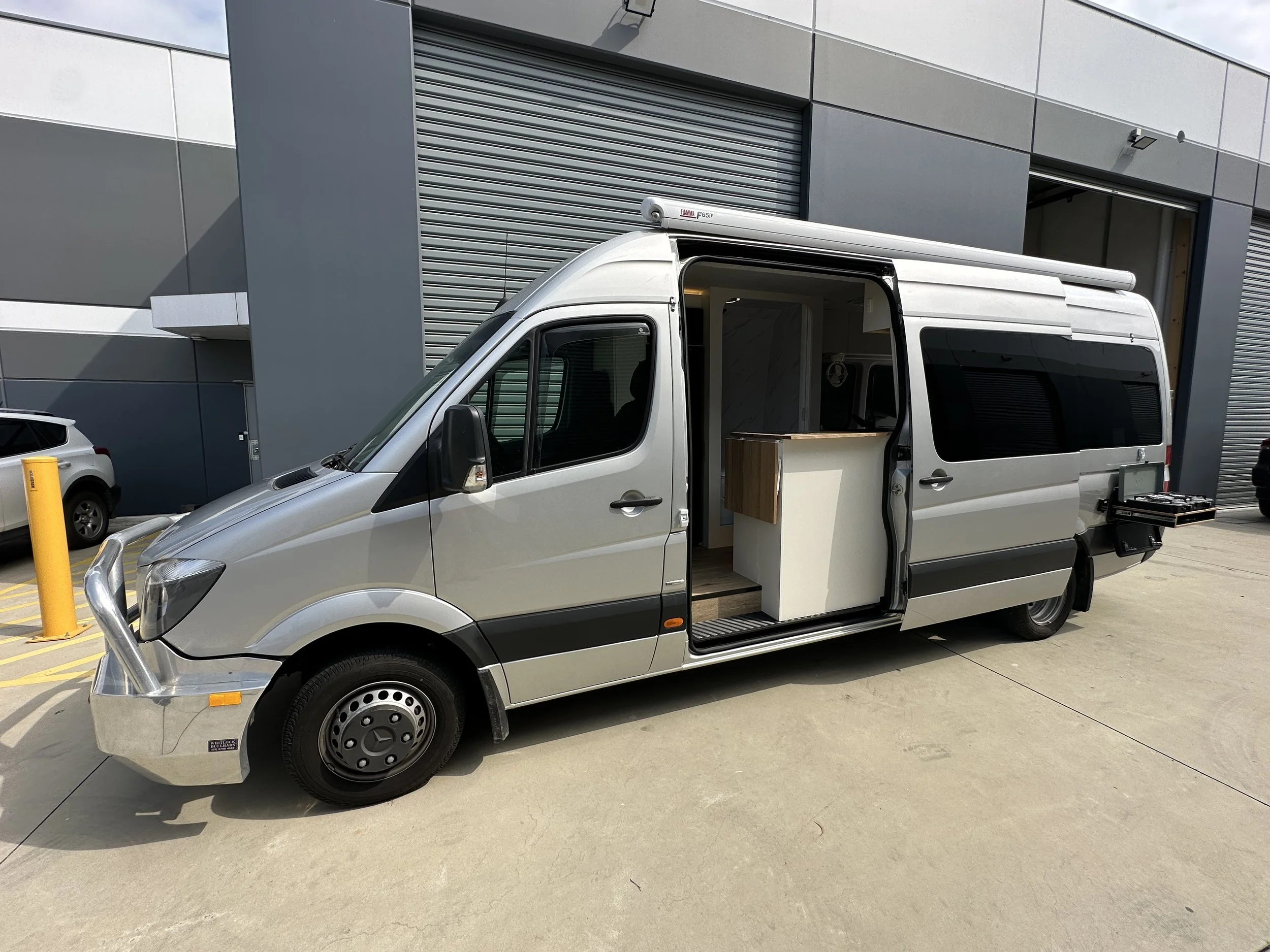 A silver camper van with an open side door revealing a small kitchen inside, parked outside a modern industrial building.