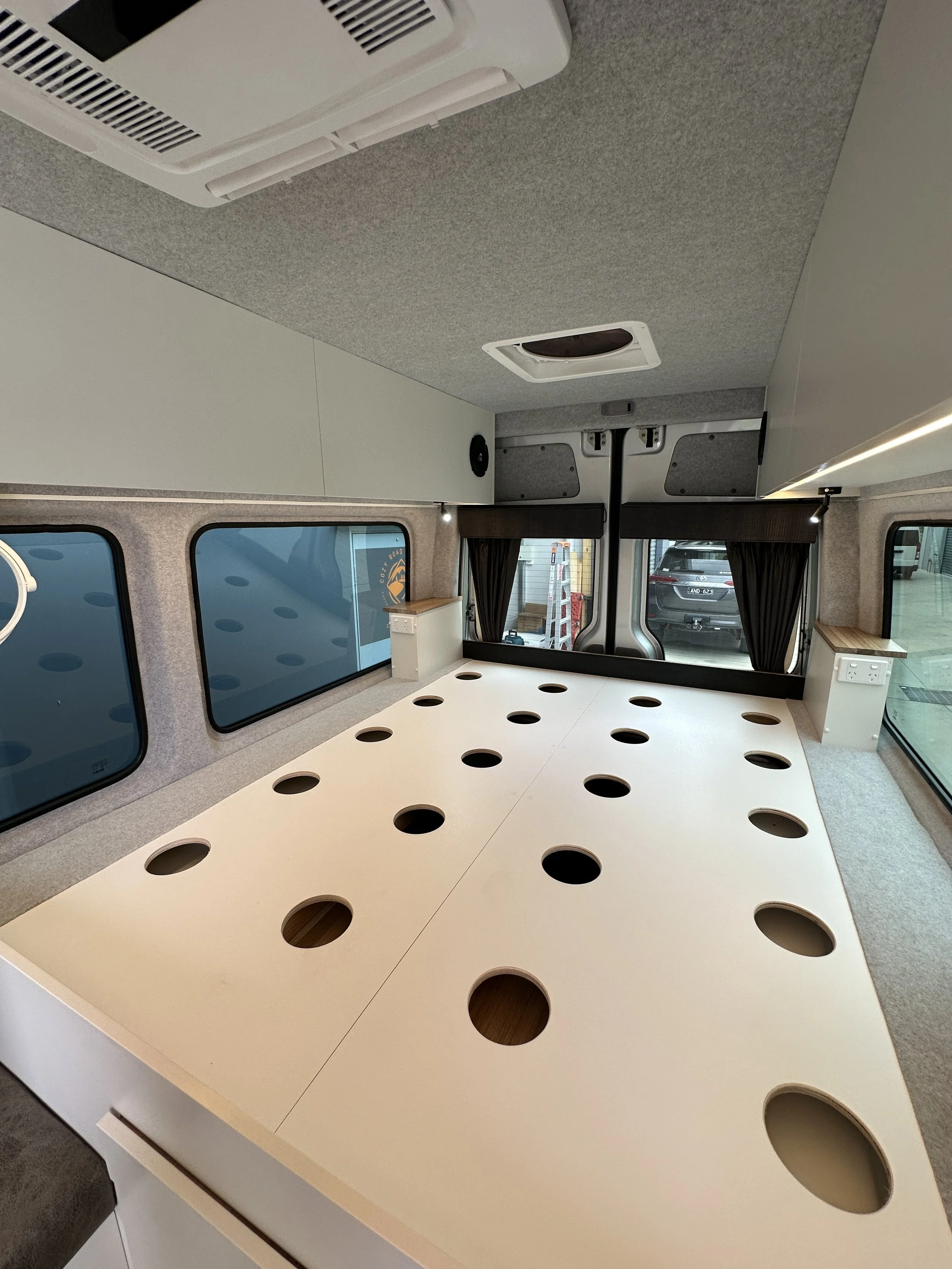 Inside a camper van or tiny house with a white table with multiple circular cutouts, side windows, and small shelves near the windows.