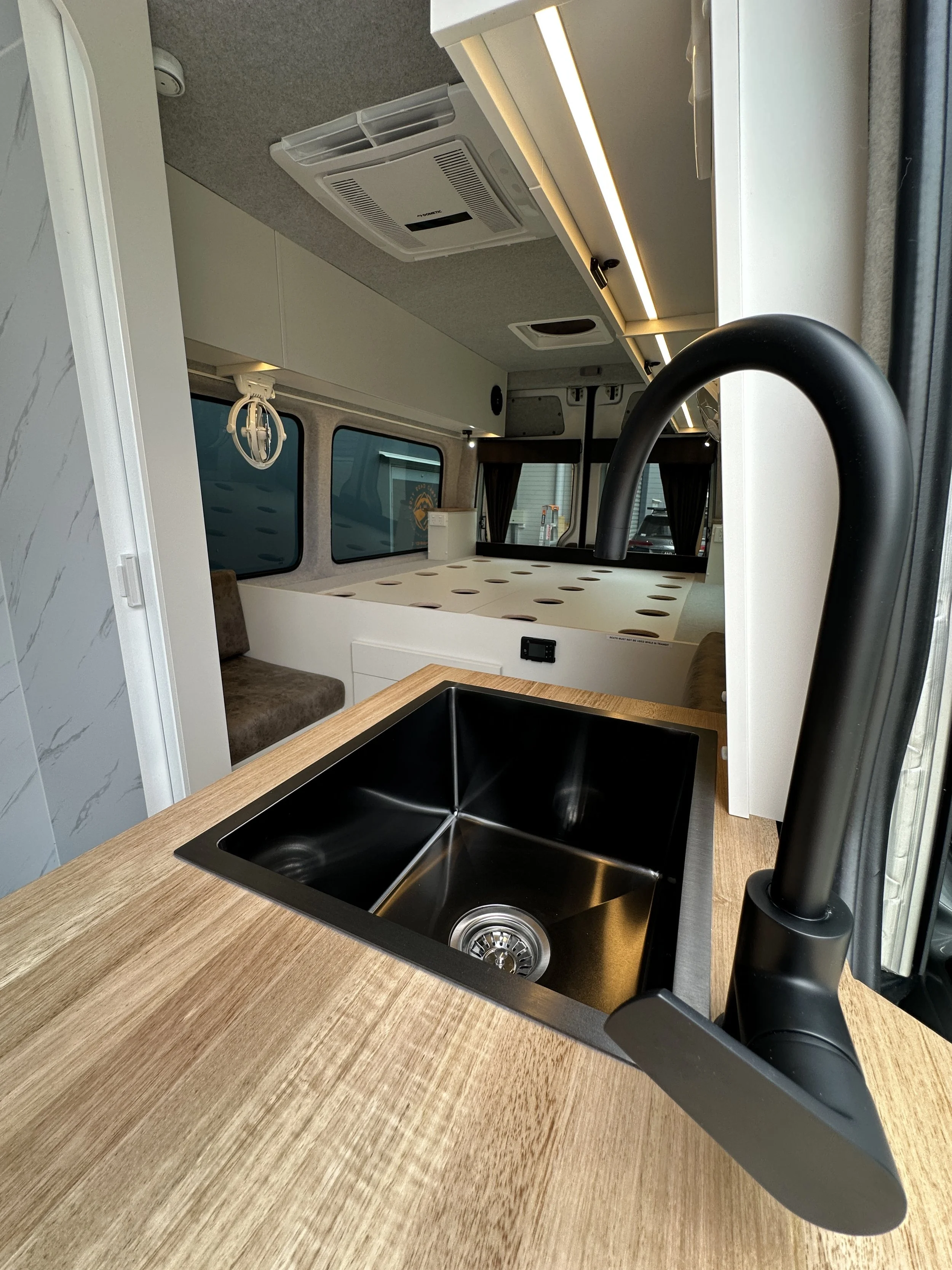Interior of a camper van showing a black kitchen sink with a curved faucet on a light wood countertop, with a sleeping area and windows in the background.
