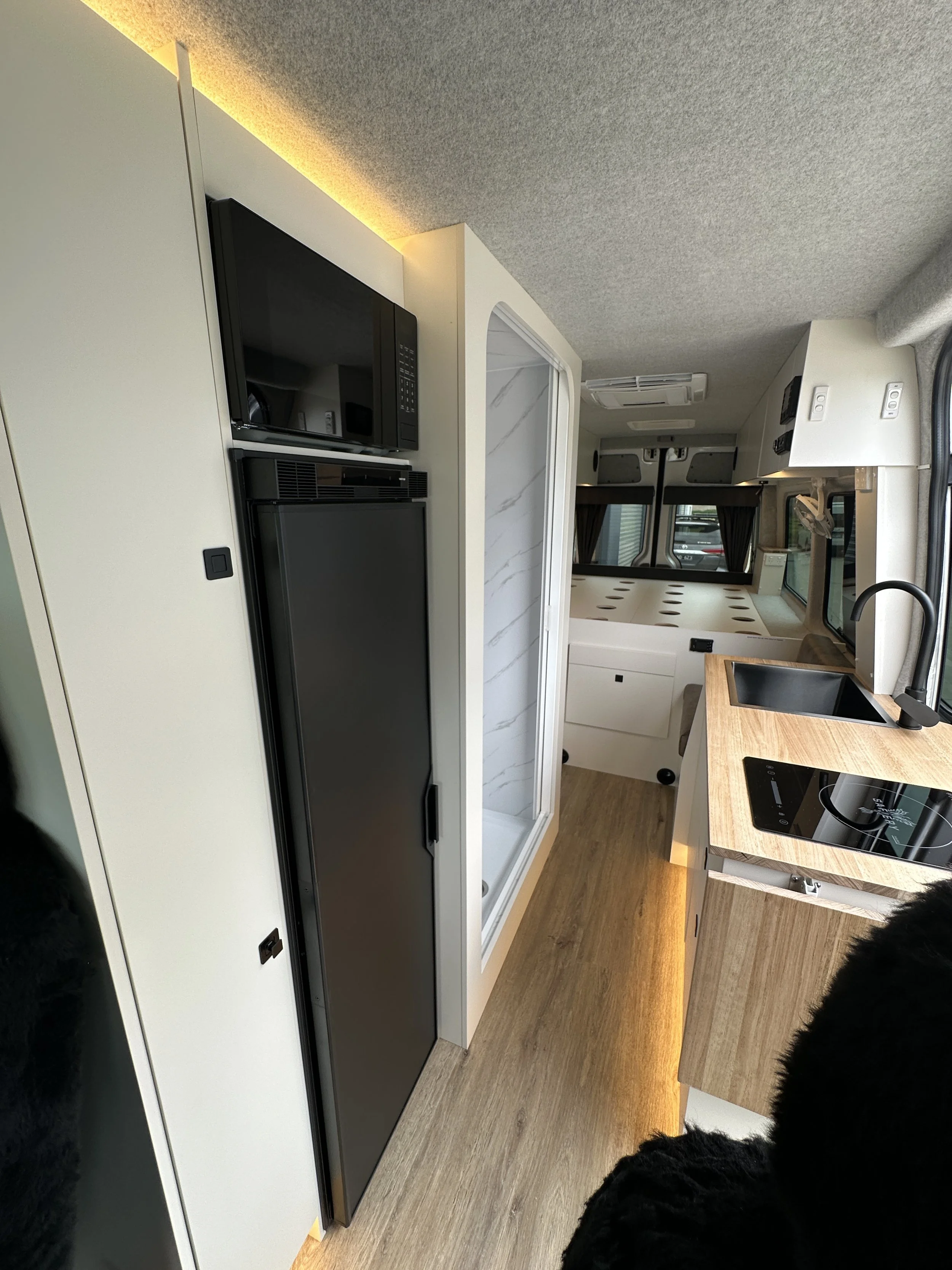 Interior of a camper van showing a microwave, a compact refrigerator, a shower stall, a countertop with a sink and electric stovetop, and a bed area with large windows and curtains.