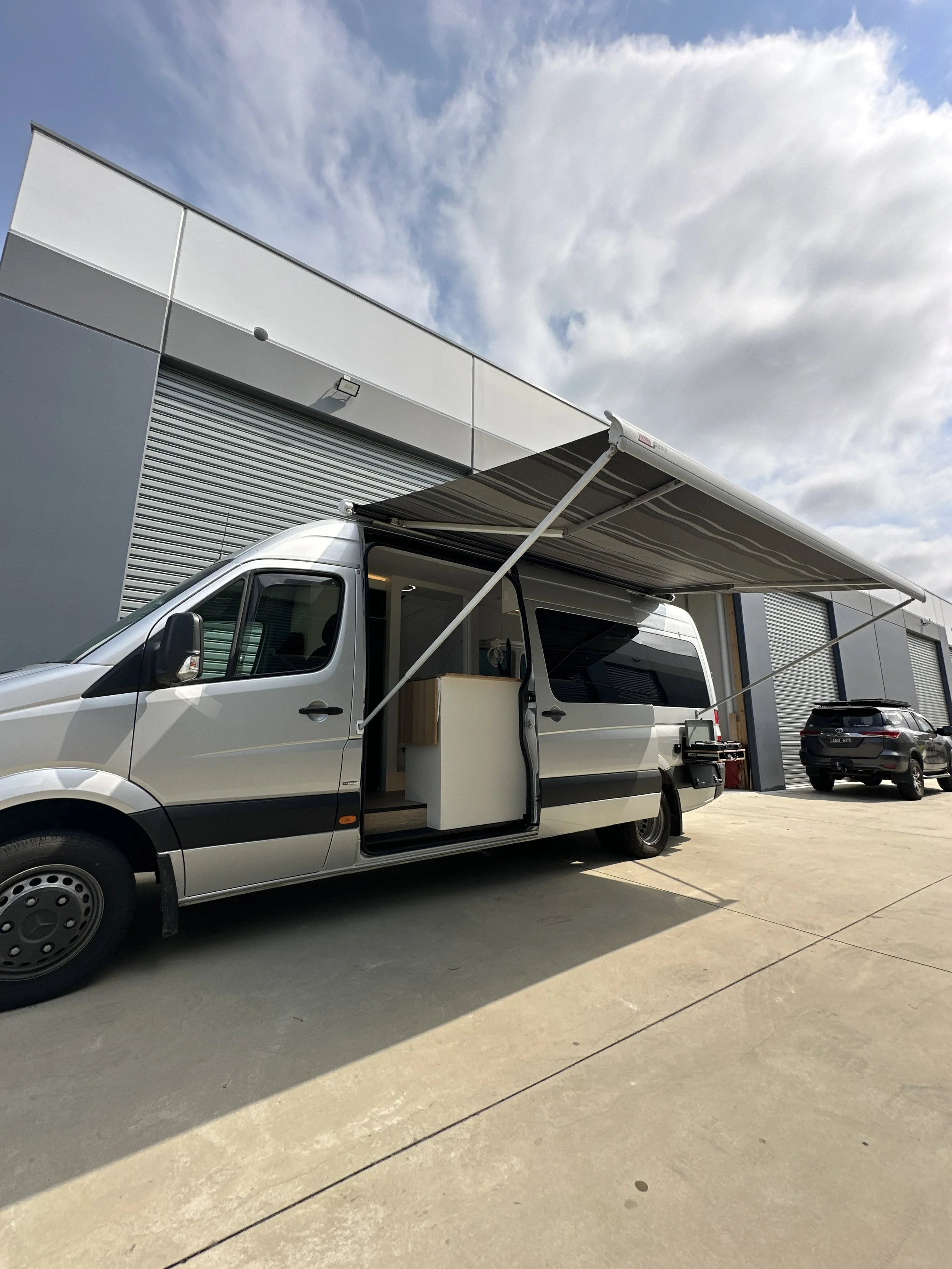 A silver van with a retractable awning extended over the side, parked outside a modern industrial building.