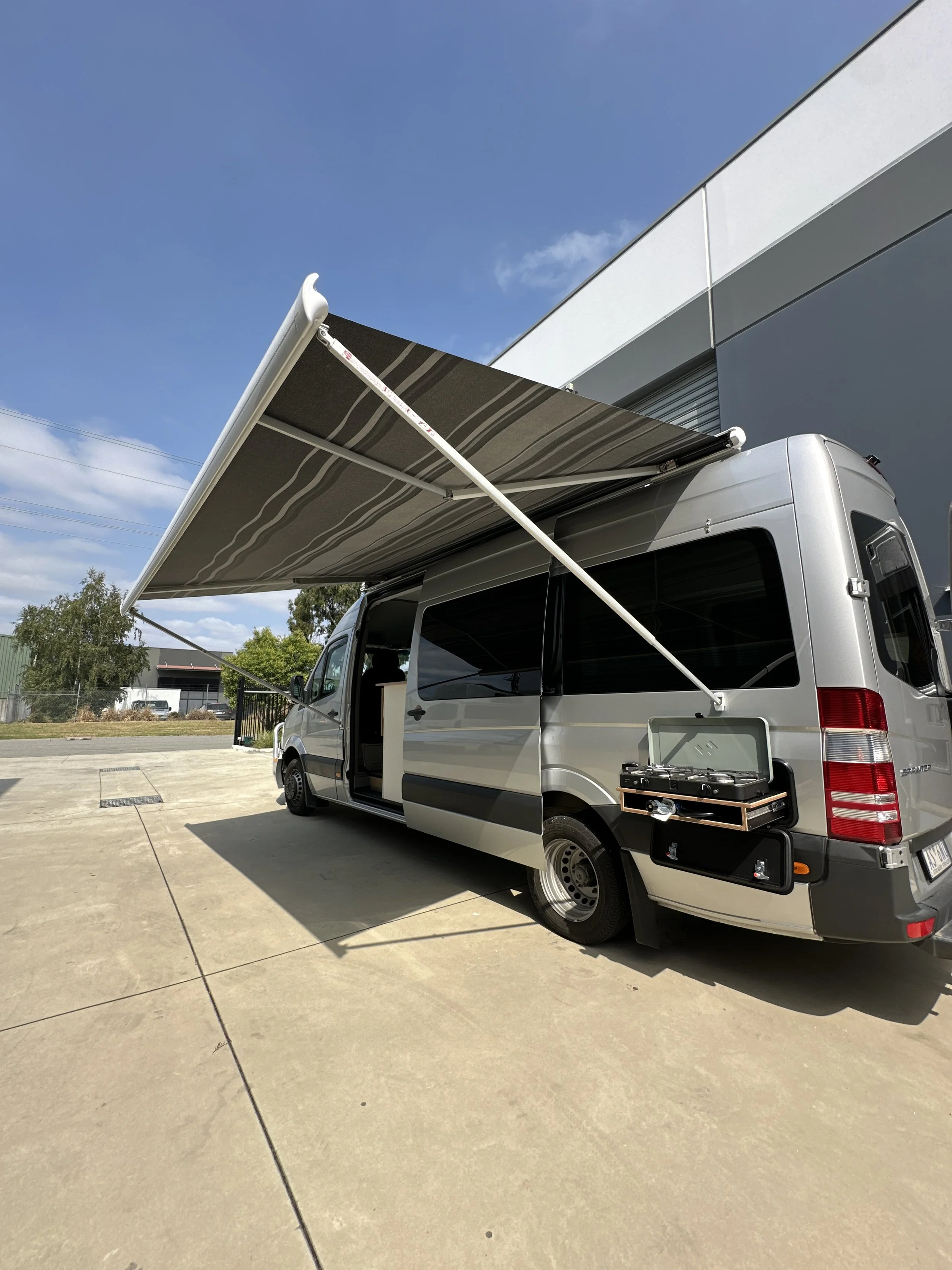 A silver camper van with a retractable awning extended on the side, parked on a concrete surface near a building under a partly cloudy sky.