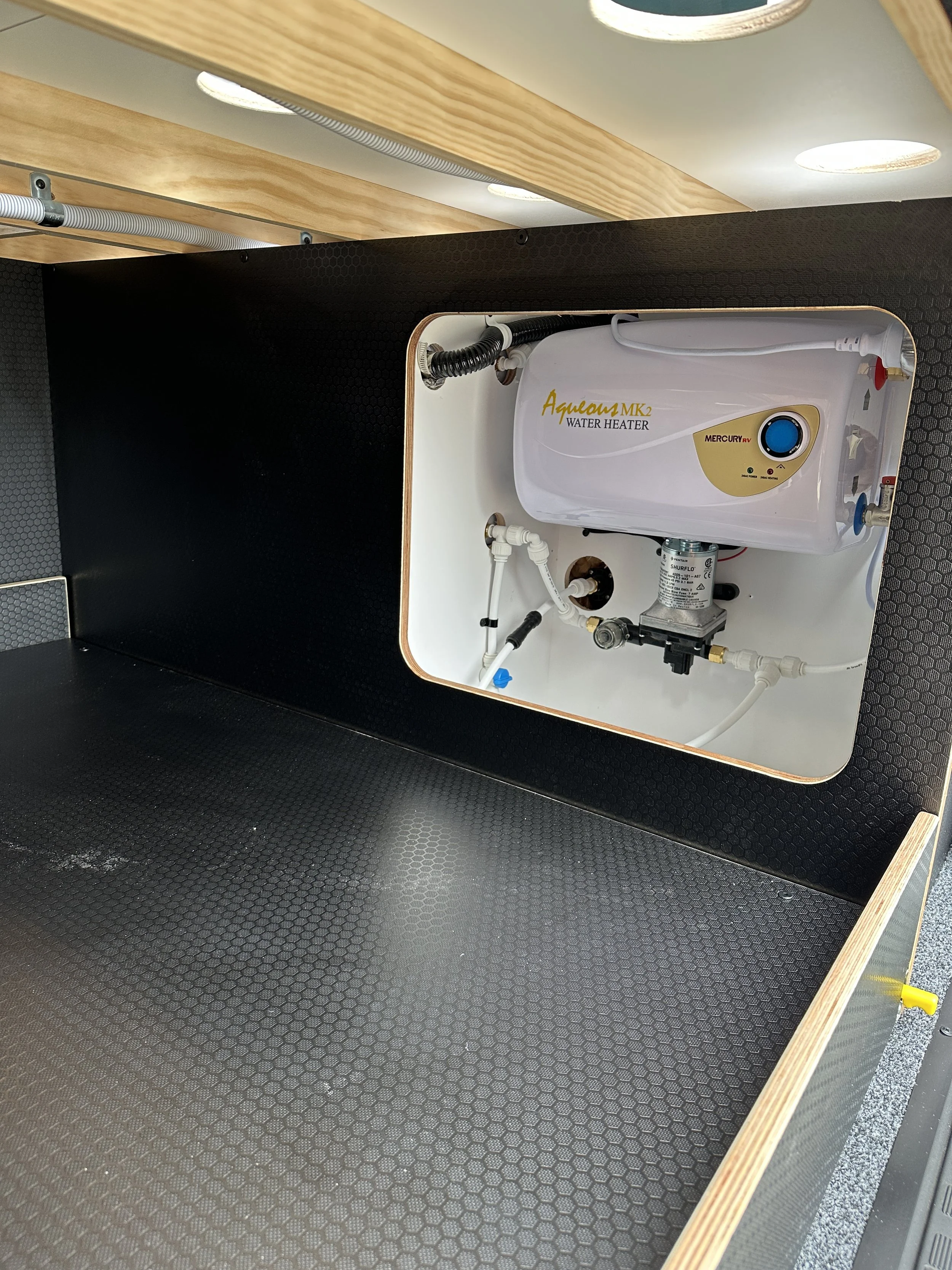 A black and white water heater inside a cubicle with black textured walls and a wooden top surface.