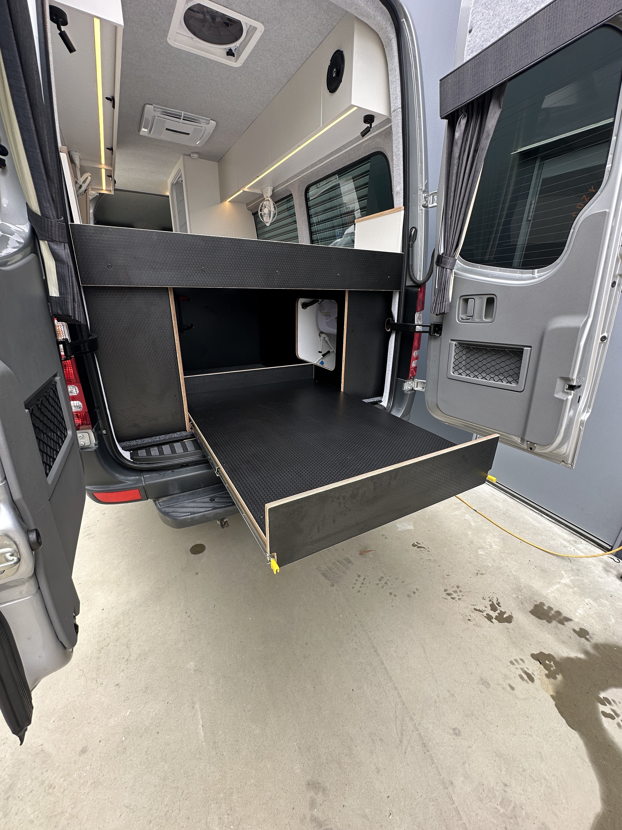 The interior of a converted camper van showing a fold-out bed platform with storage beneath and a kitchenette area inside.