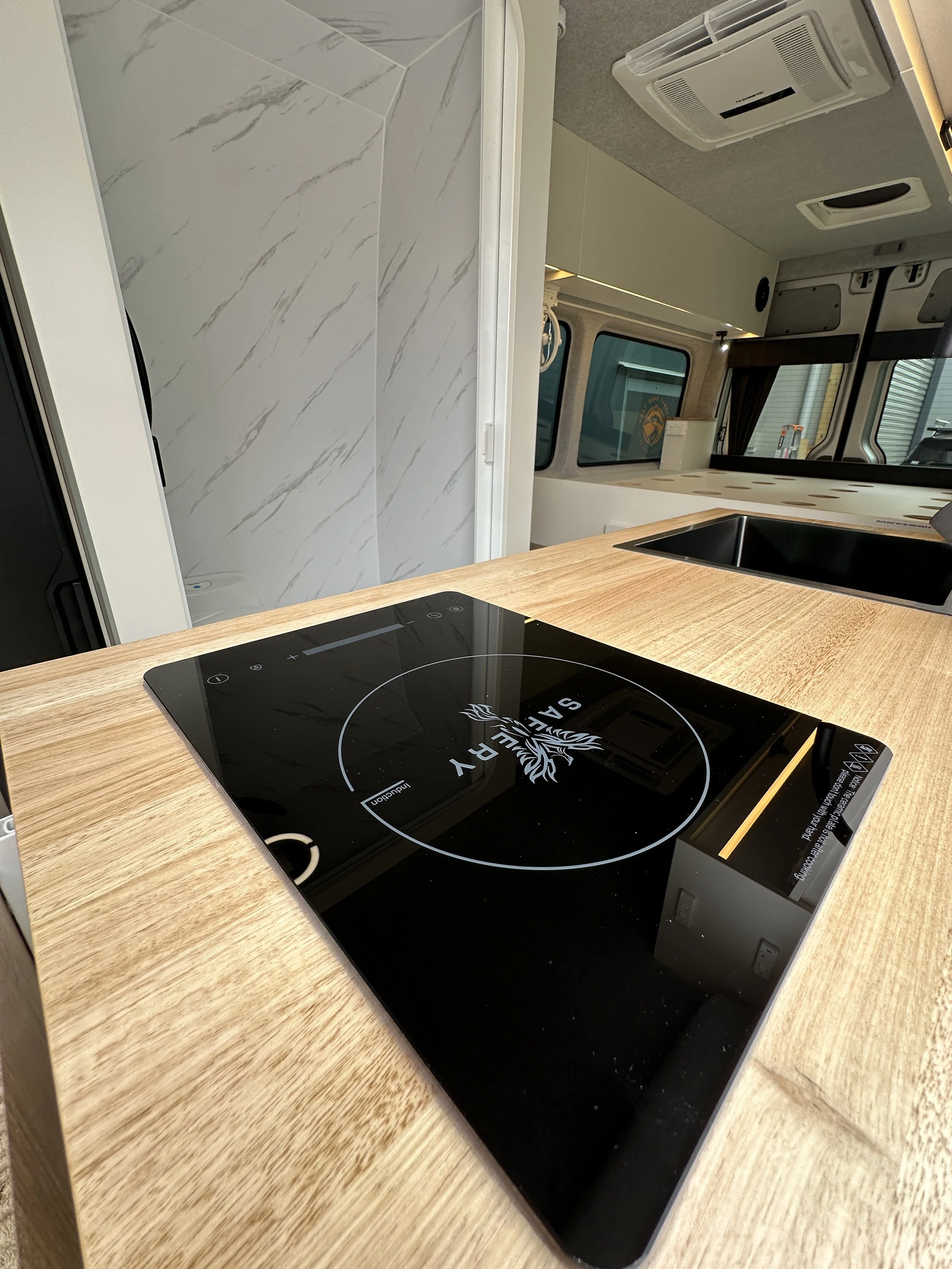 A black induction cooktop with a logo and control panel on a wooden kitchen counter inside a camper or RV.