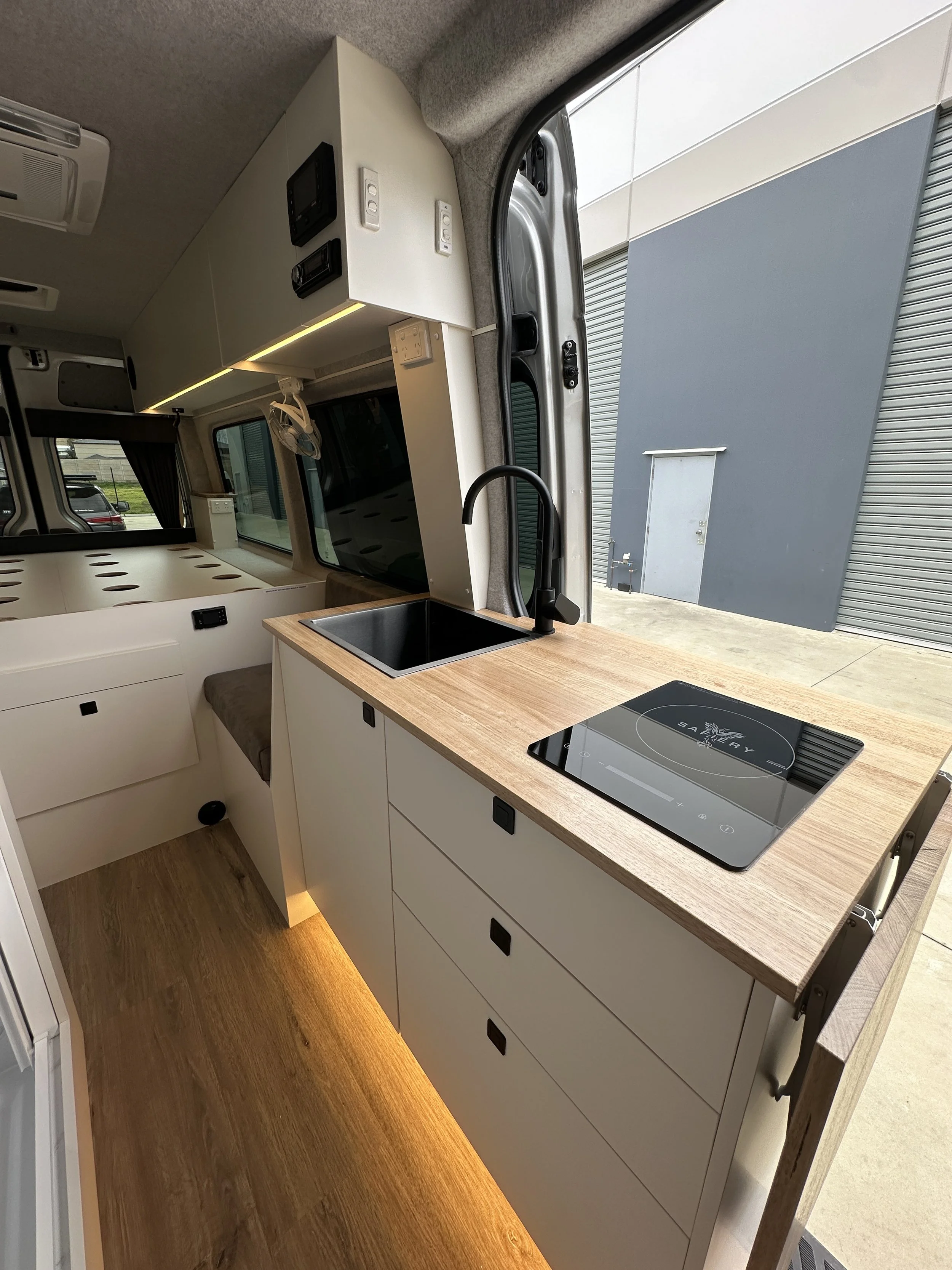 Interior of a compact camper van kitchen with a wooden countertop, black sink, and electric cooktop, large window, and storage cabinets.